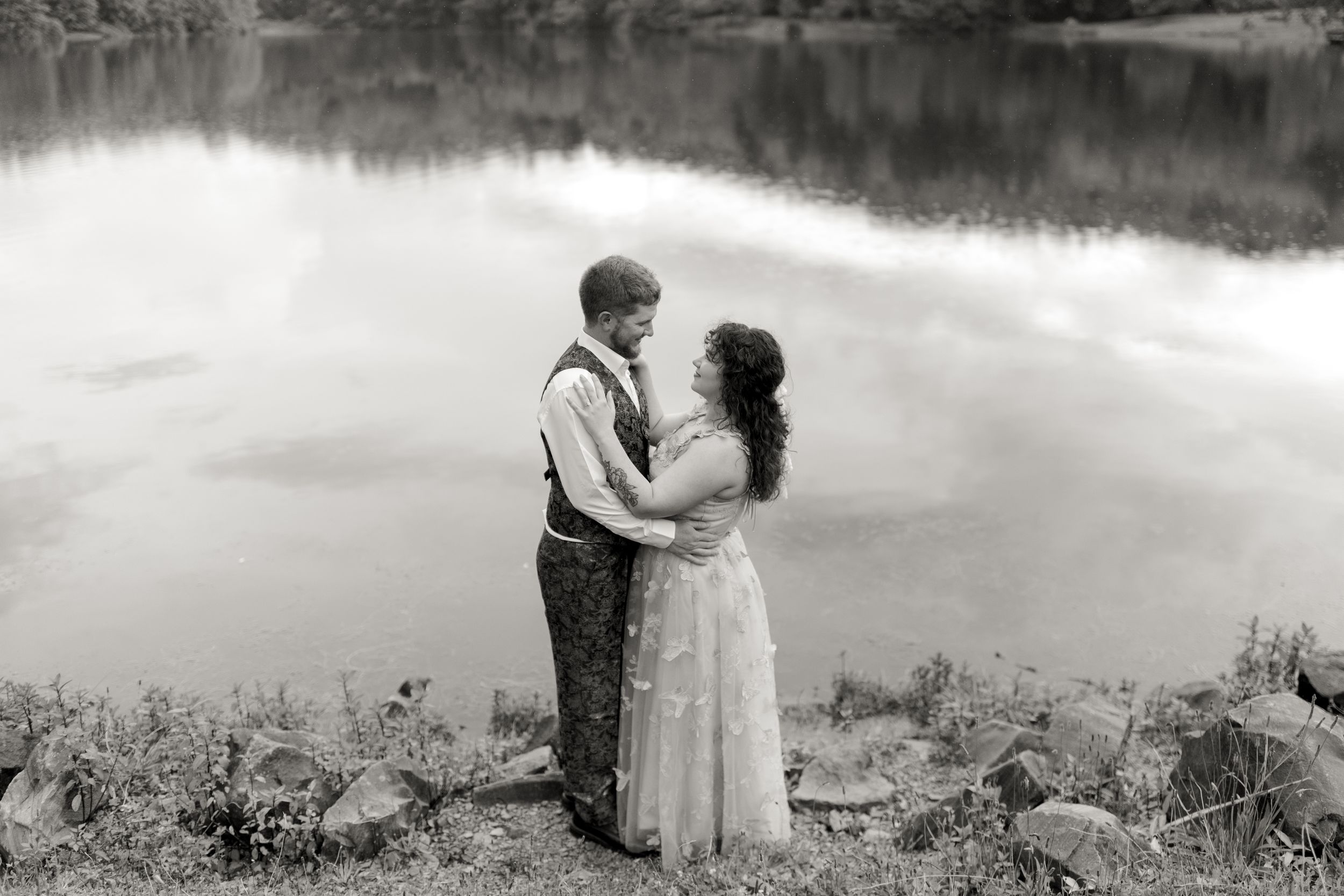 A couple embraces by a tranquil lake on a cloudy day, with rocks and vegetation along the shore.