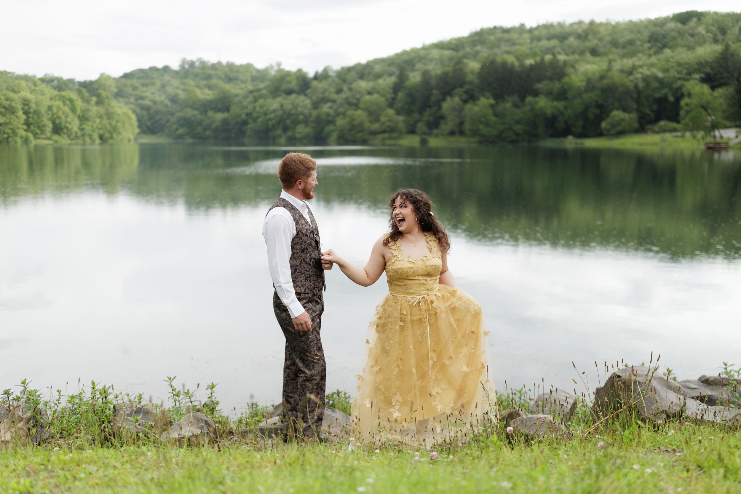 A couple holds hands by a serene lake with green hills in the background, woman wearing a sparkly gold gown.