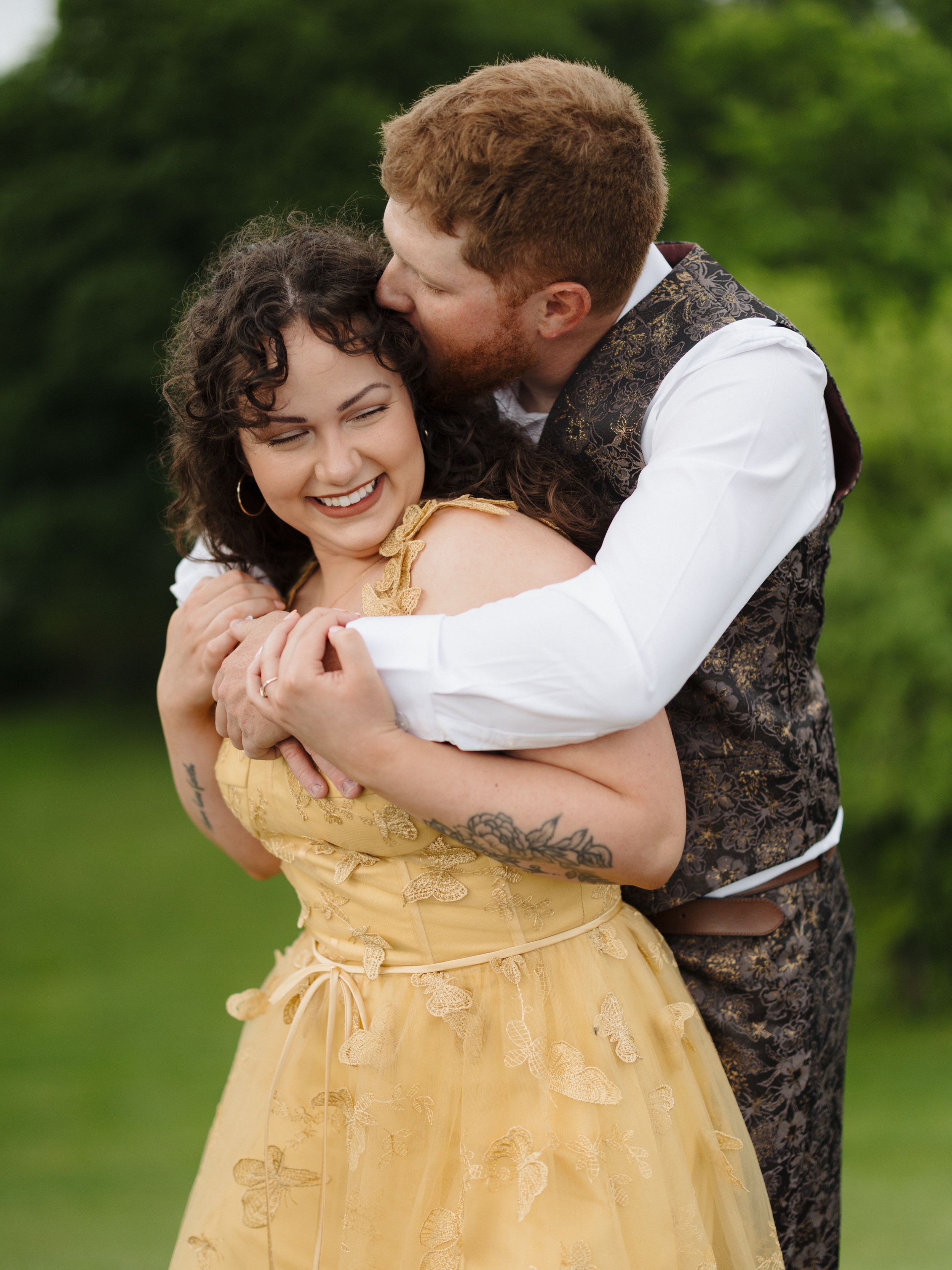 A joyful couple embraces outdoors, with her wearing a yellow dress and him in a patterned vest and white shirt.