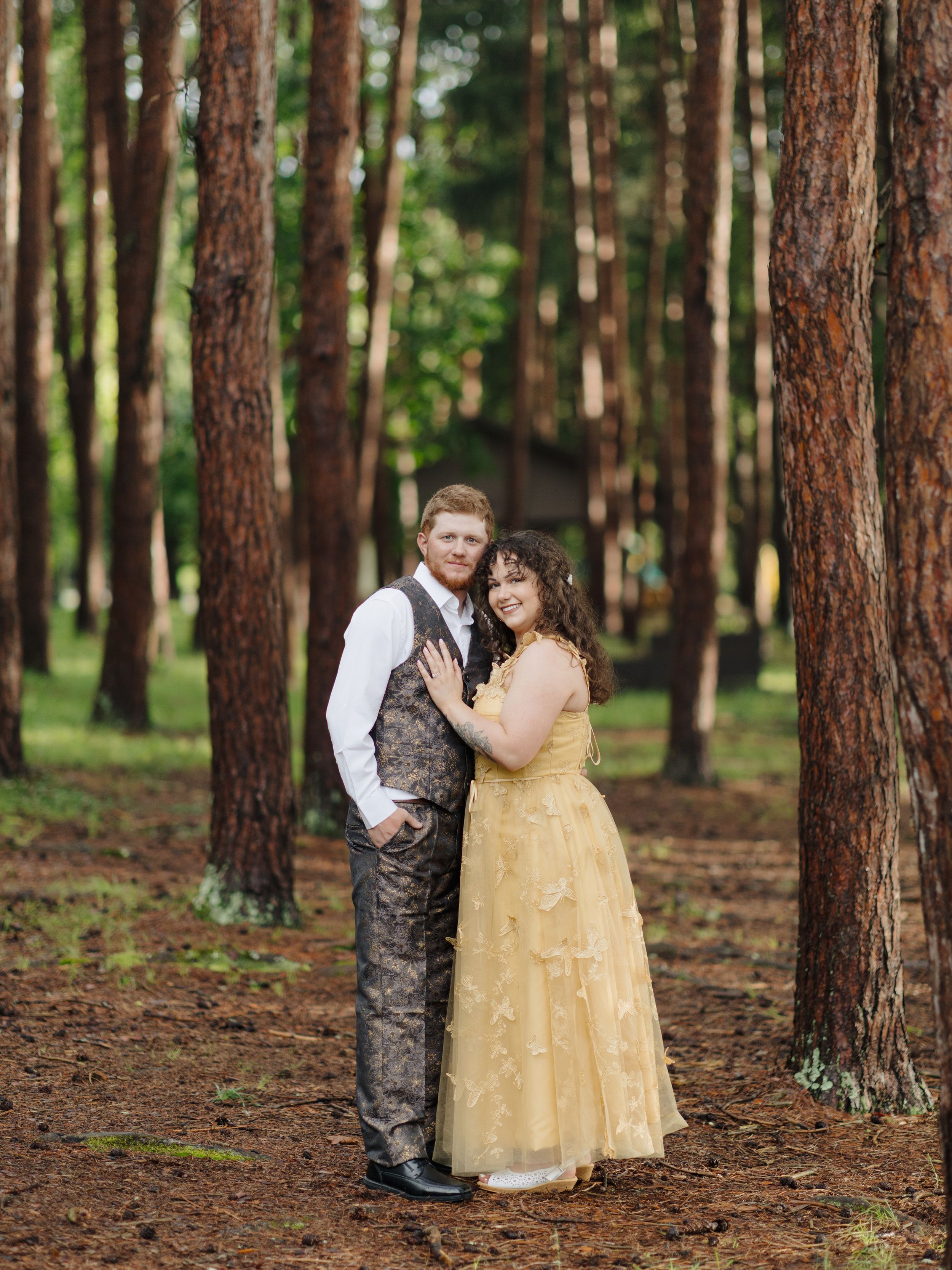 A couple in formal wear embraces in a pine forest, with the woman in gold and man in brown vest and white shirt.
