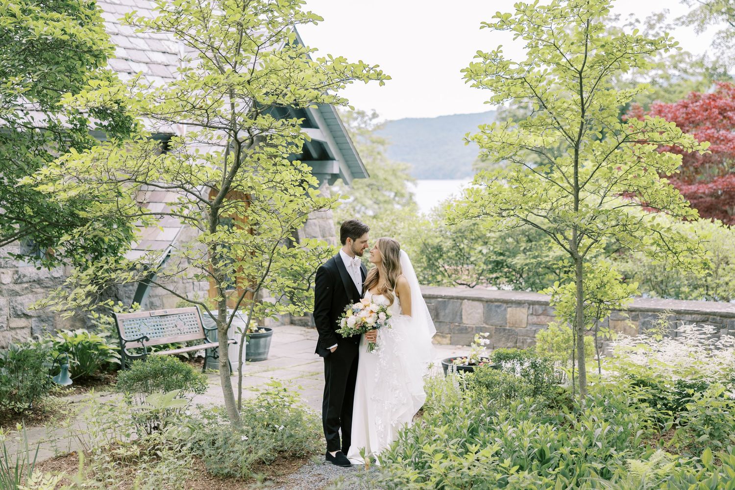 A couple stand together on a garden path by the Silver Bay YMCA chapel surrounded by lush green trees and foliage and Lake George.