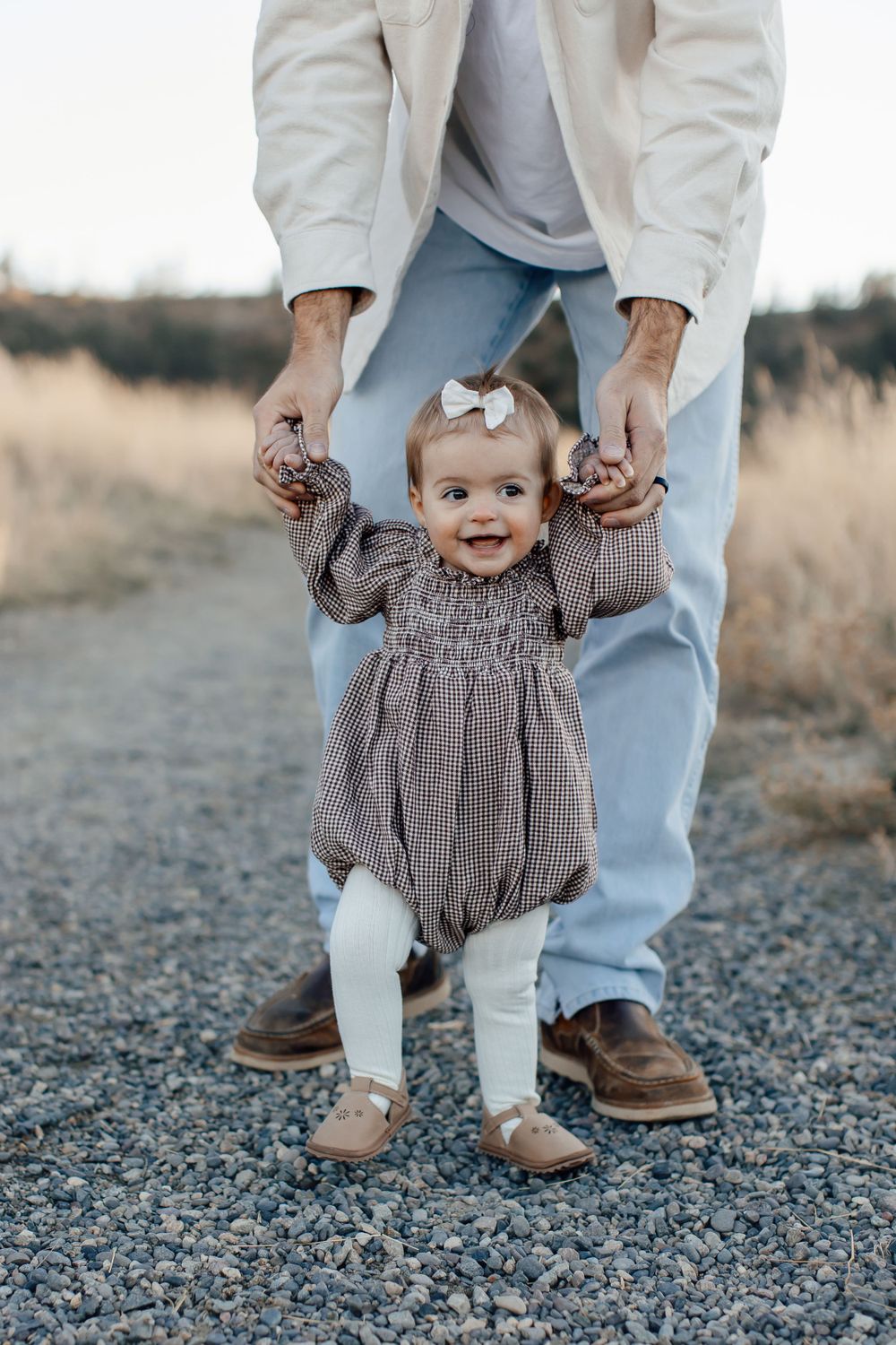 Small child in checkered dress smiles while learning to walk on gravel path.