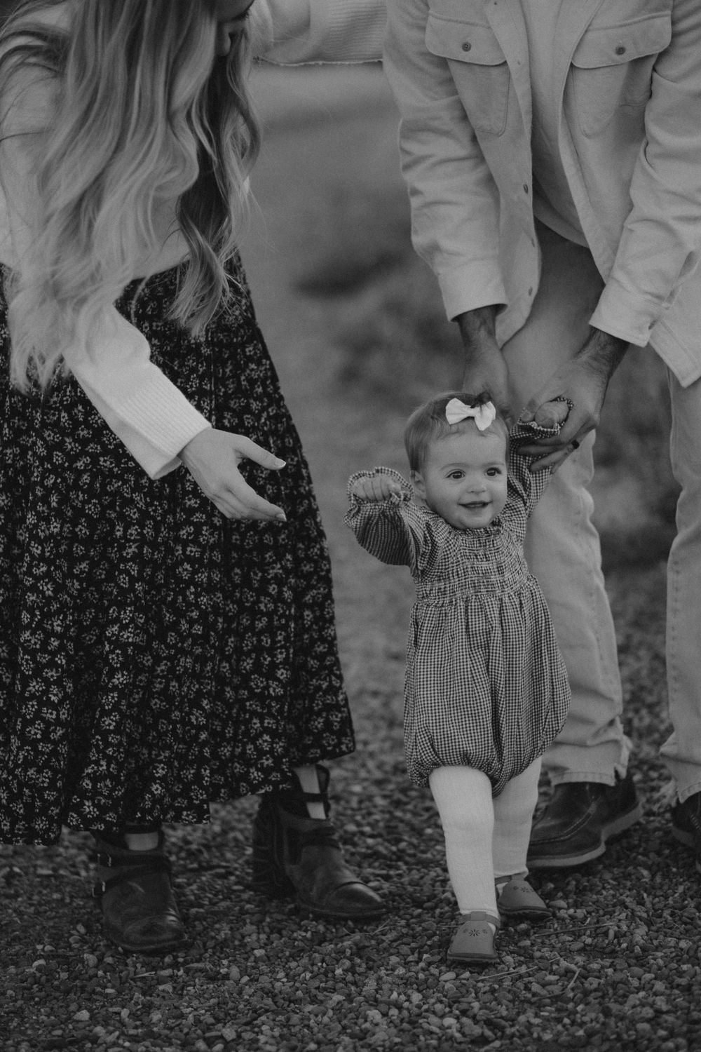 A toddler in a patterned dress walks while holding hands with adults on either side.