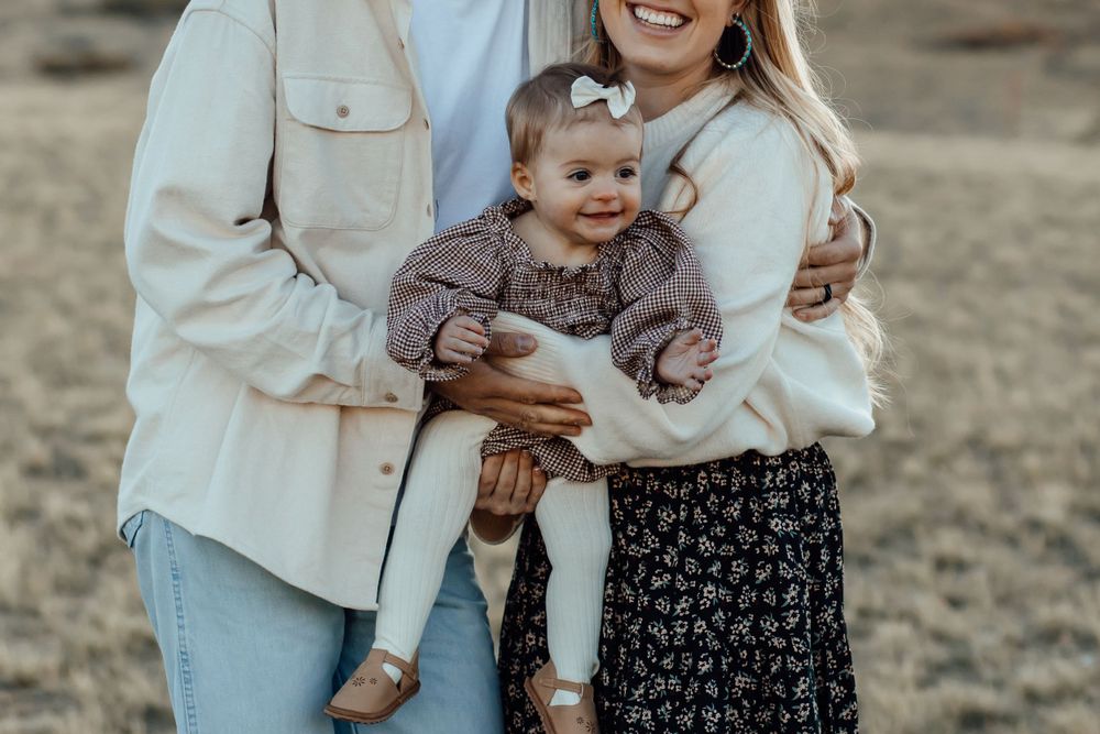 A couple holds their baby while posing in casual attire against a natural landscape.