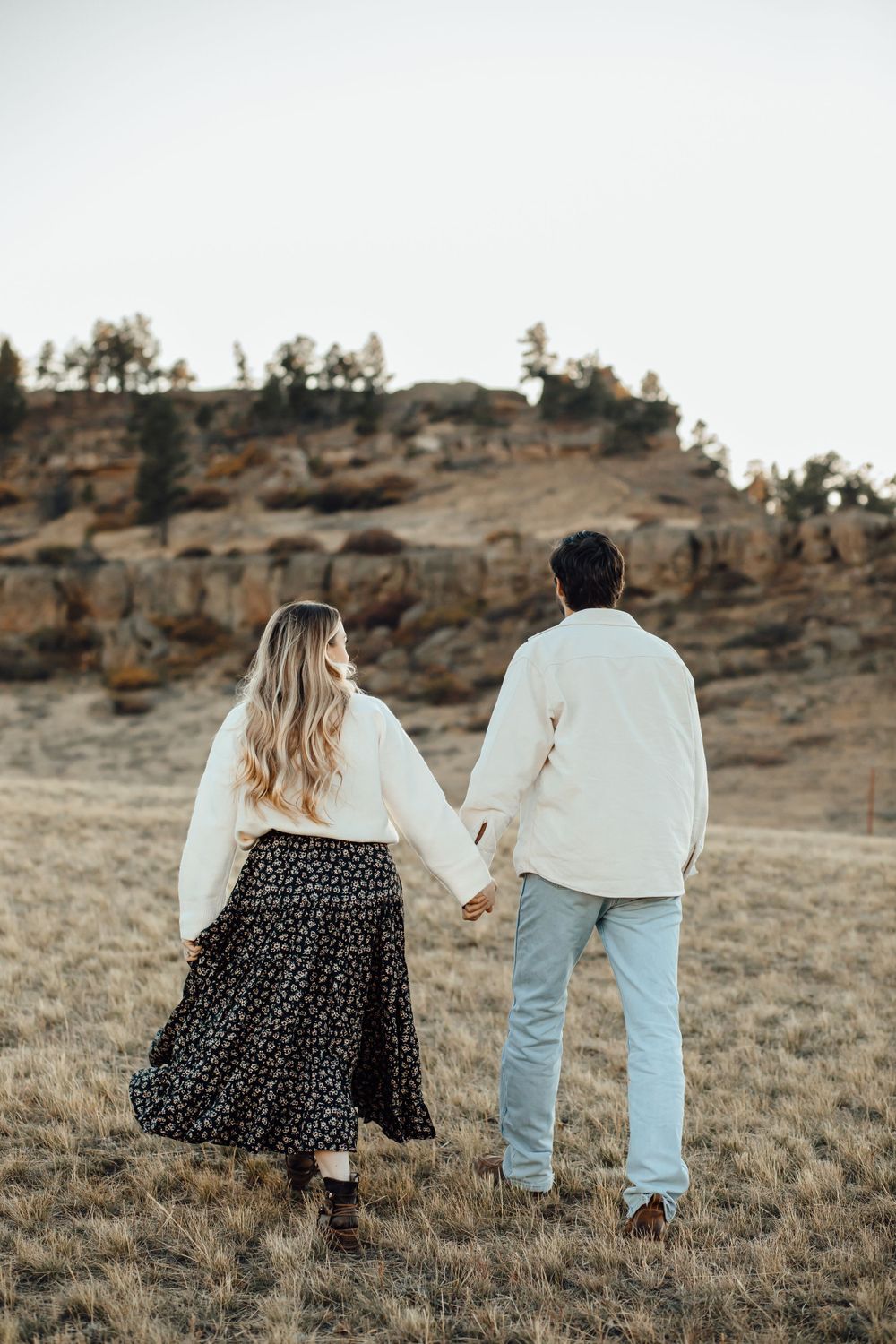 A couple in matching white sweaters holds hands while walking in a mountainous field.