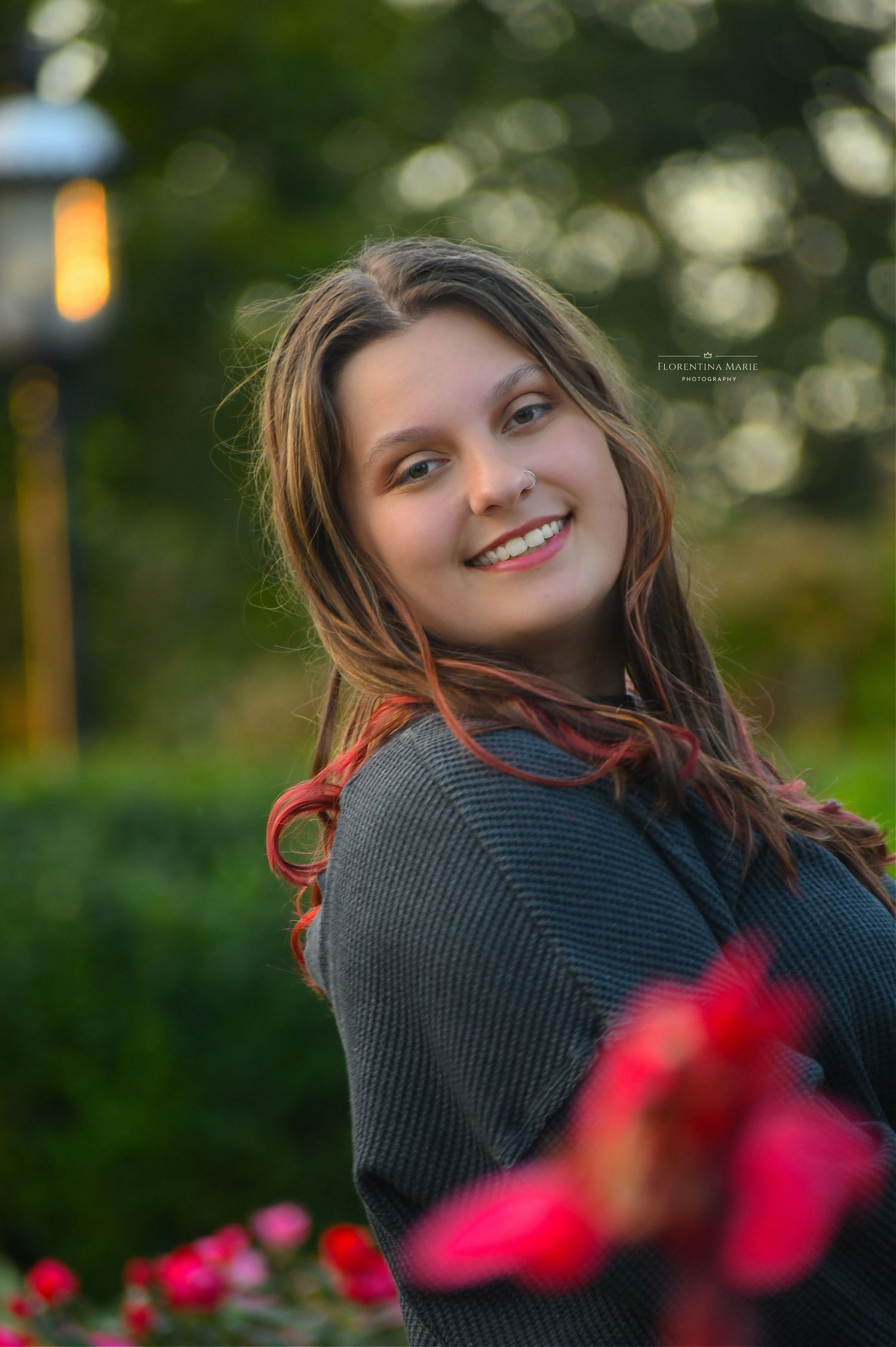 Outdoor portrait of a young woman in a dark sweater smiling near red flowers at twilight.