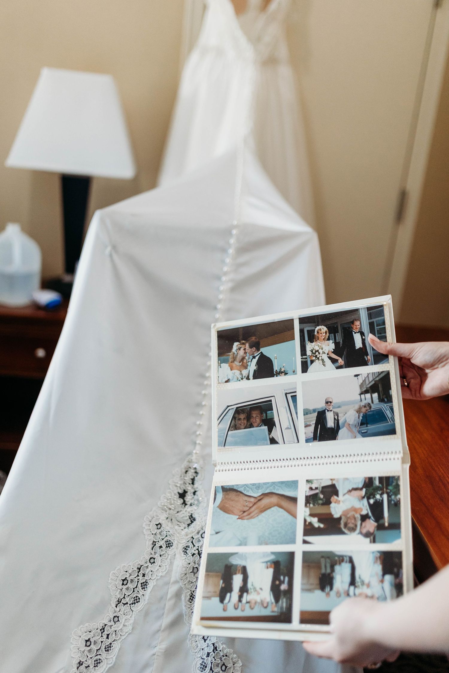 Person holding wedding photo album in front of a lace bridal gown hanging in a room.