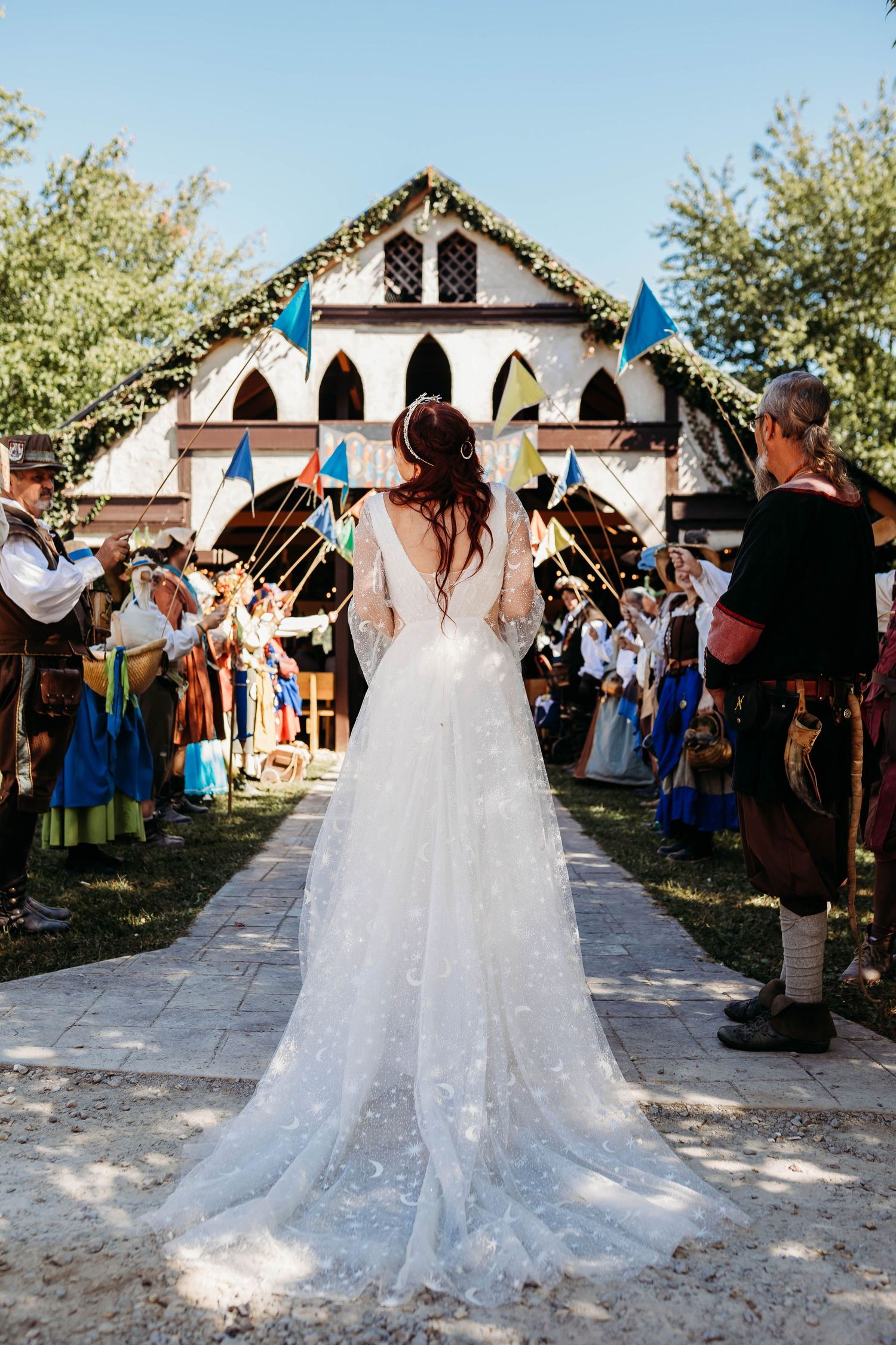 Bride in floral lace gown walks toward medieval chapel at Renaissance faire wedding ceremony.