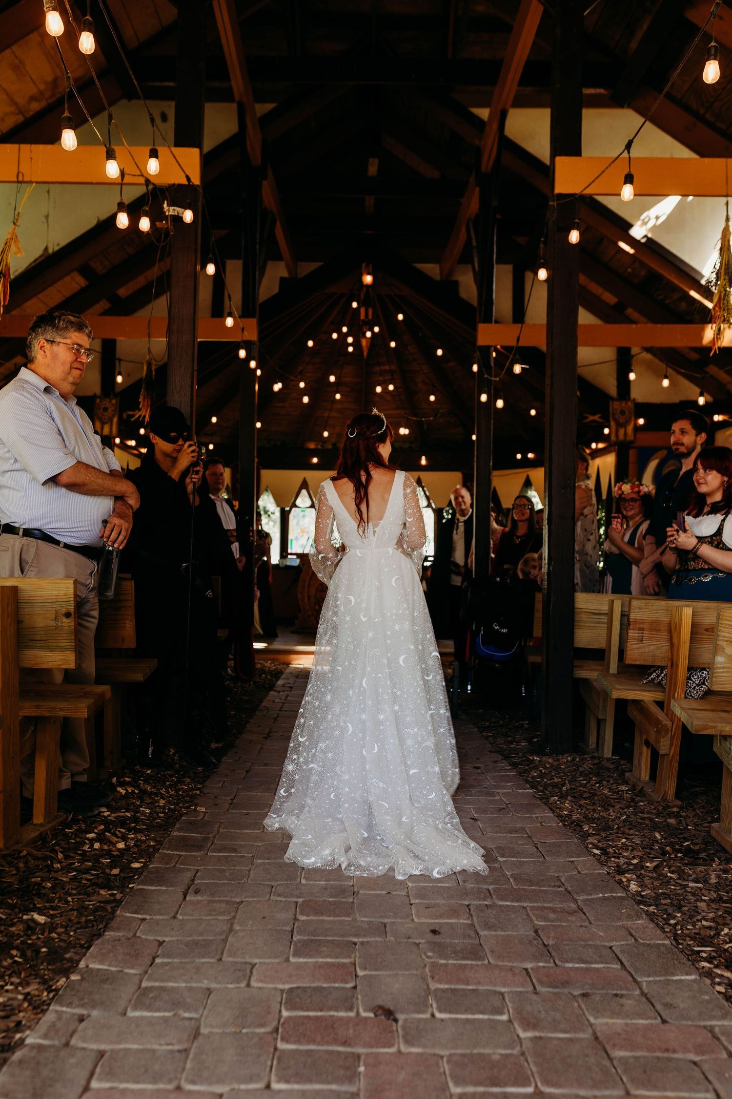 Bride in floral tulle gown walks down rustic barn aisle with string lights overhead.