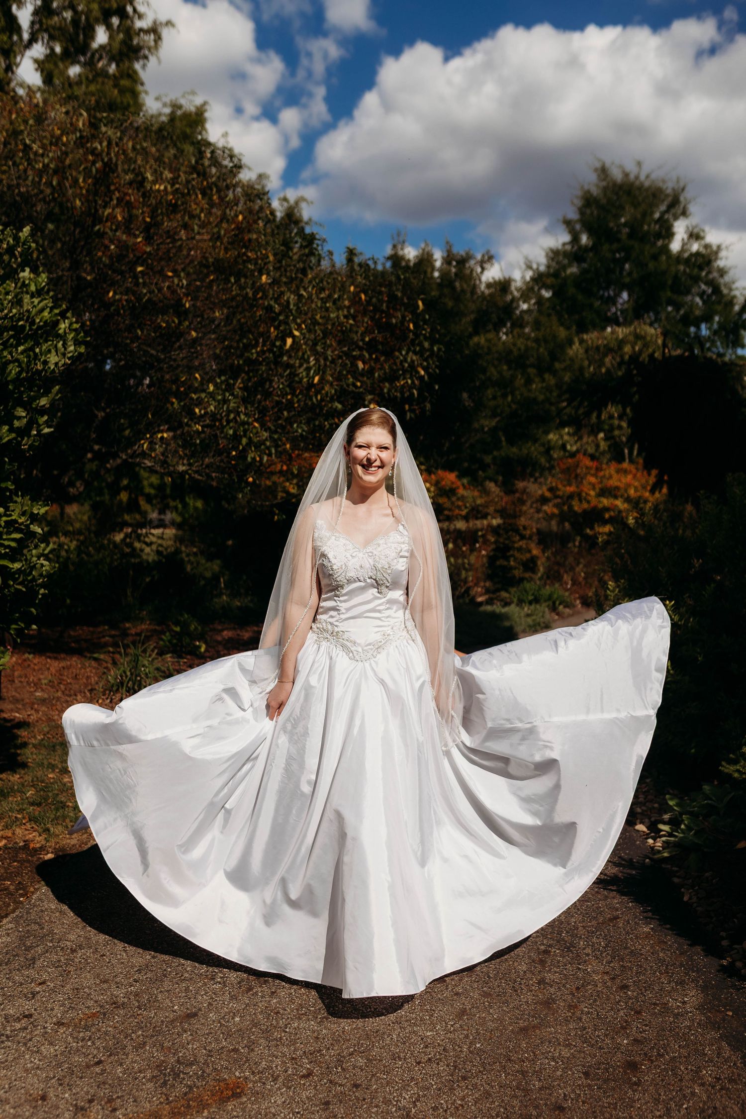 Smiling bride in white ballgown and veil twirling in a sunlit garden under blue sky.