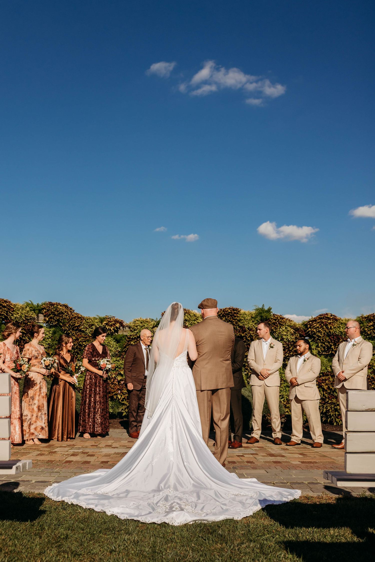 Bride in white gown with cathedral train at outdoor wedding ceremony under blue sky with wedding party.