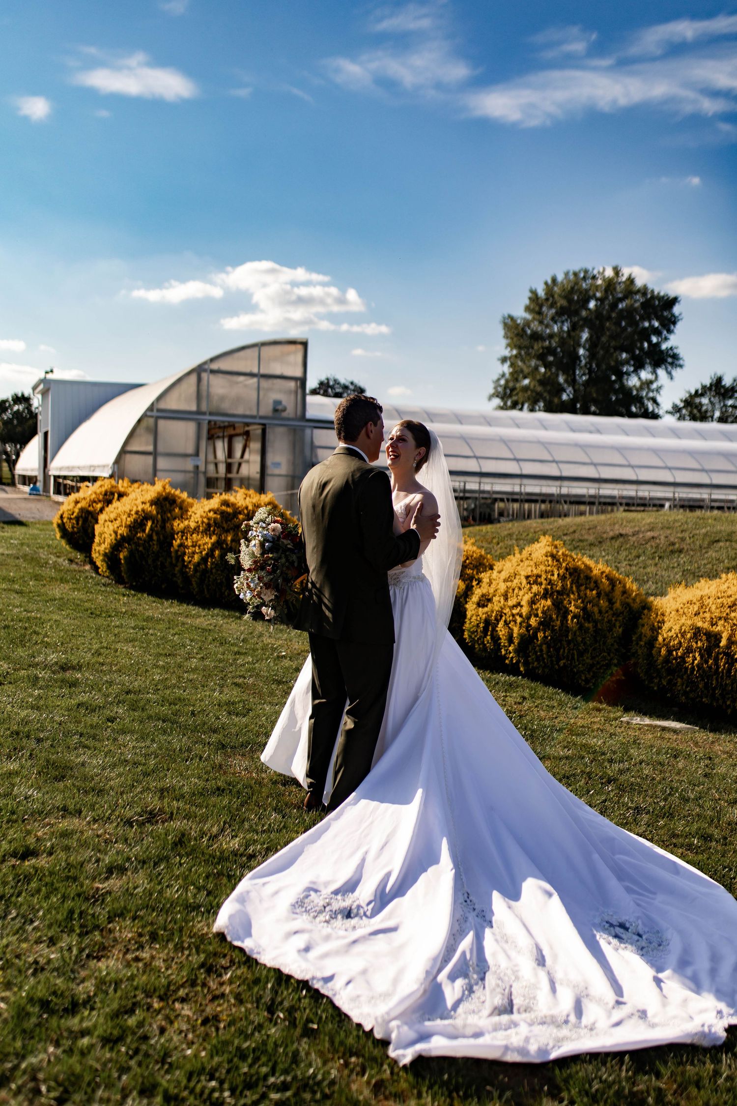 Bride and groom embrace on green lawn with long bridal train spread out near greenhouse venue.