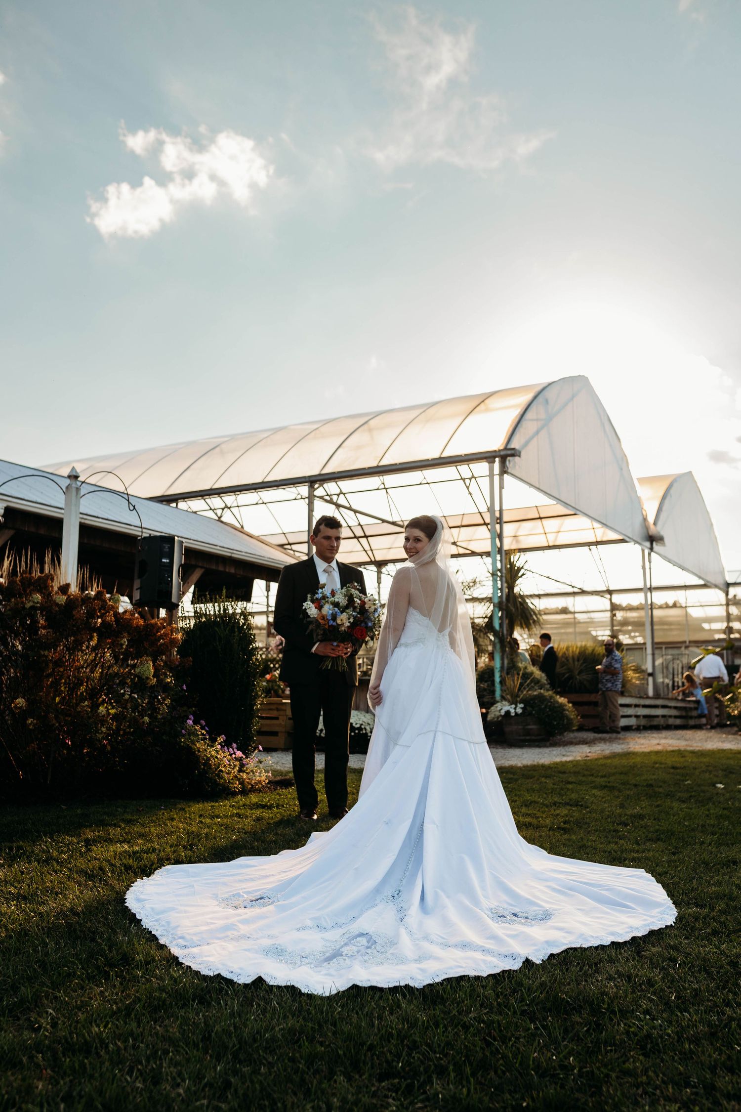 Bride in backless gown with long train stands with groom near greenhouse wedding venue at sunset.