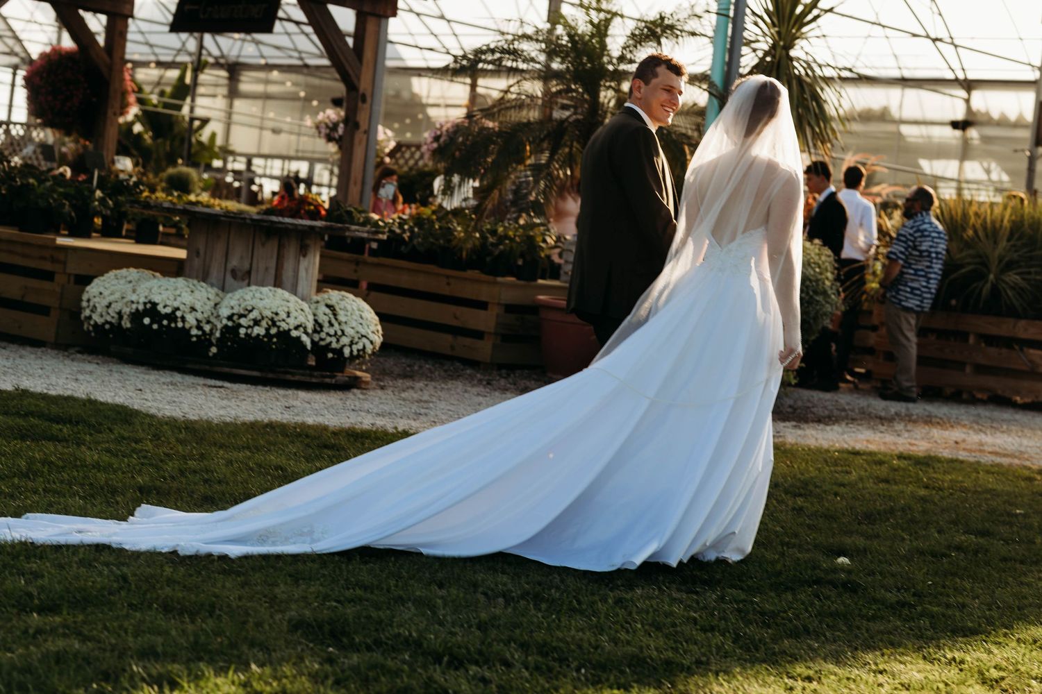 Bride and groom walk together near greenhouse venue with elegant white cathedral train trailing on grass.
