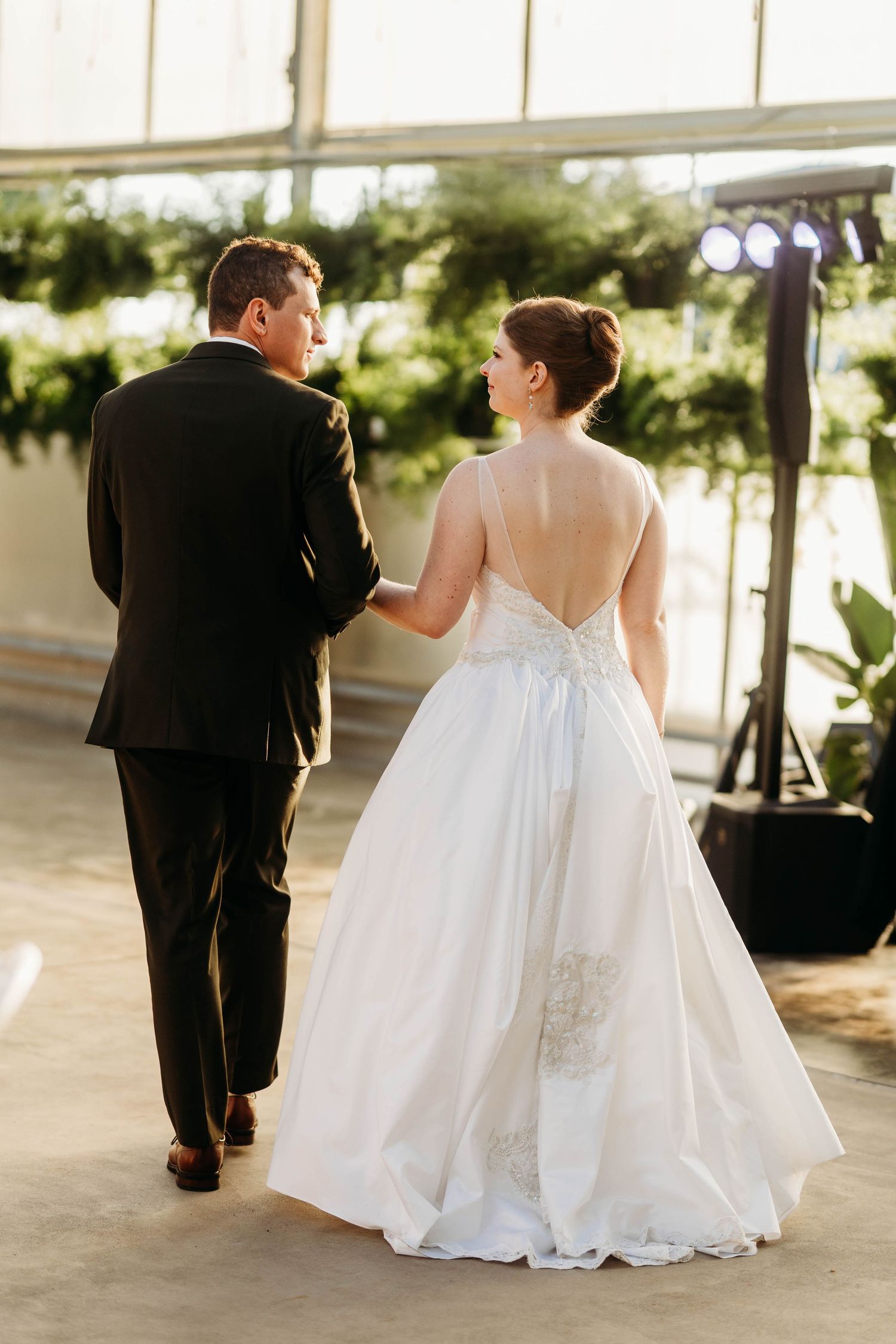 Bride in backless lace wedding gown walks hand in hand with groom through greenhouse reception venue.