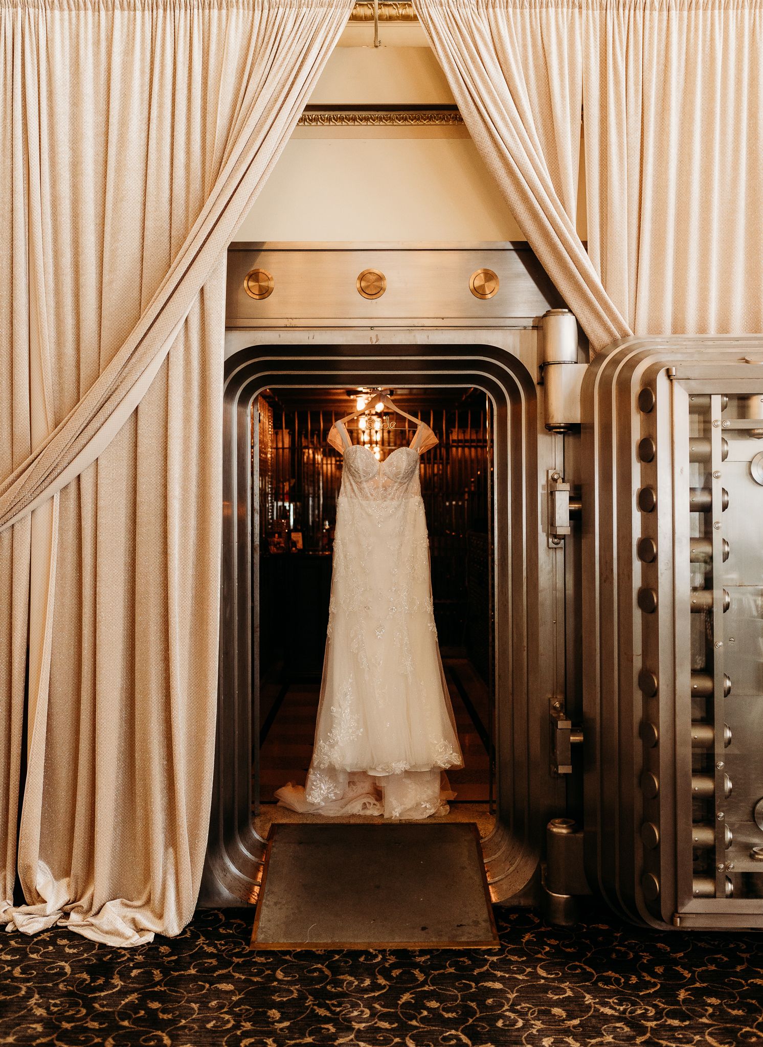 Elegant lace wedding dress displayed in ornate bank vault doorway with draped curtains.