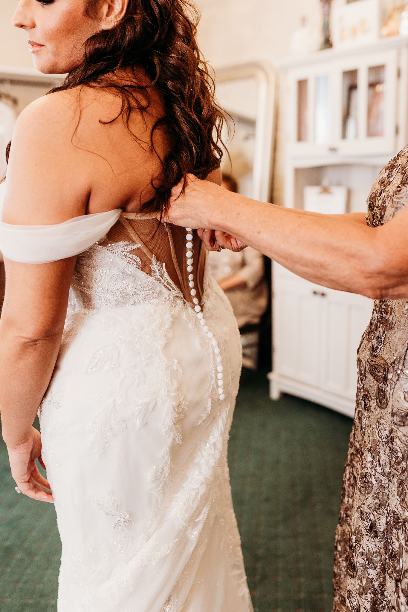 Mother fastening pearl button back closure on brides lace off-shoulder wedding gown.