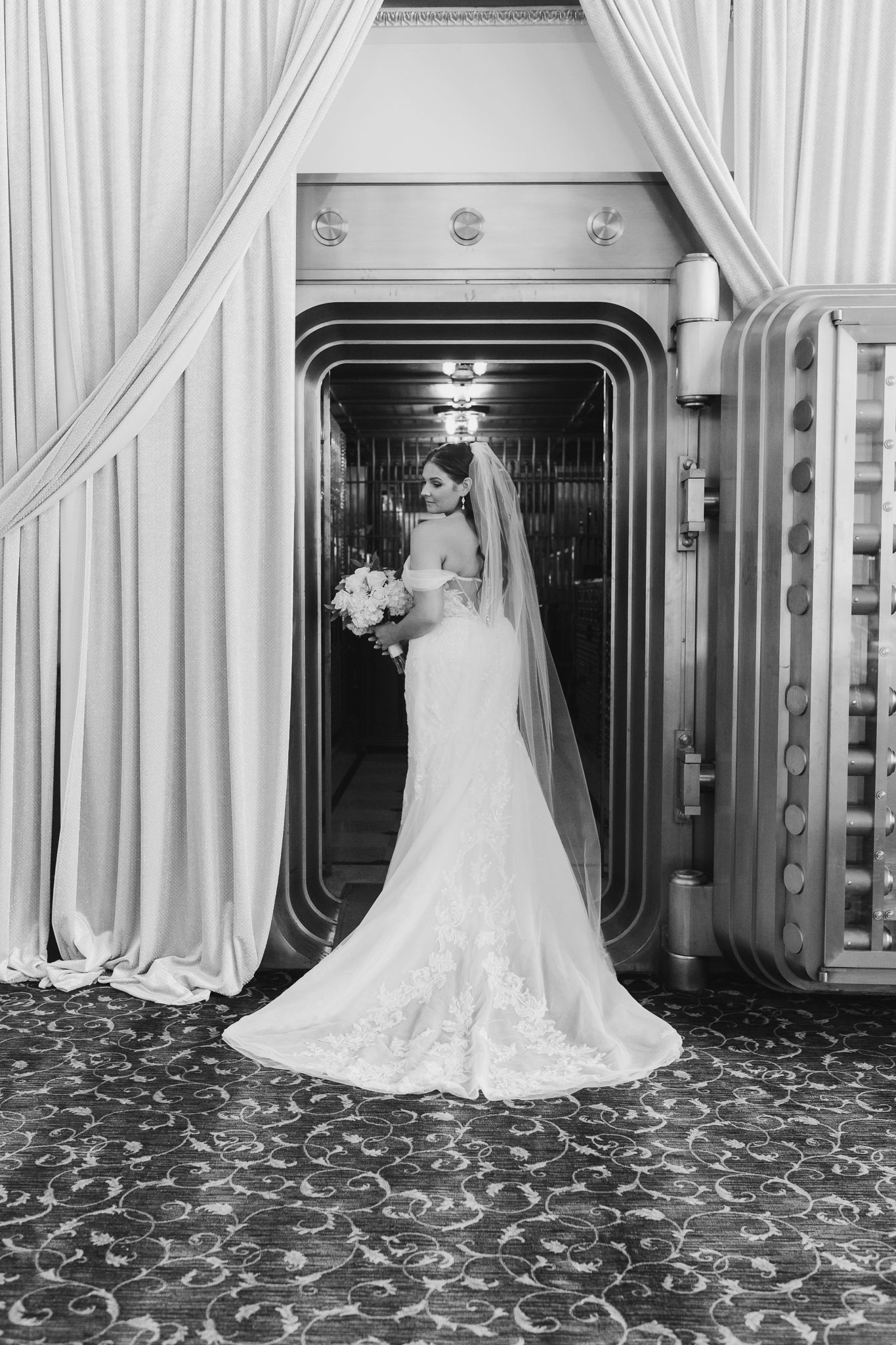 Bride in lace mermaid gown holding bouquet poses in vintage bank vault doorway in black and white.