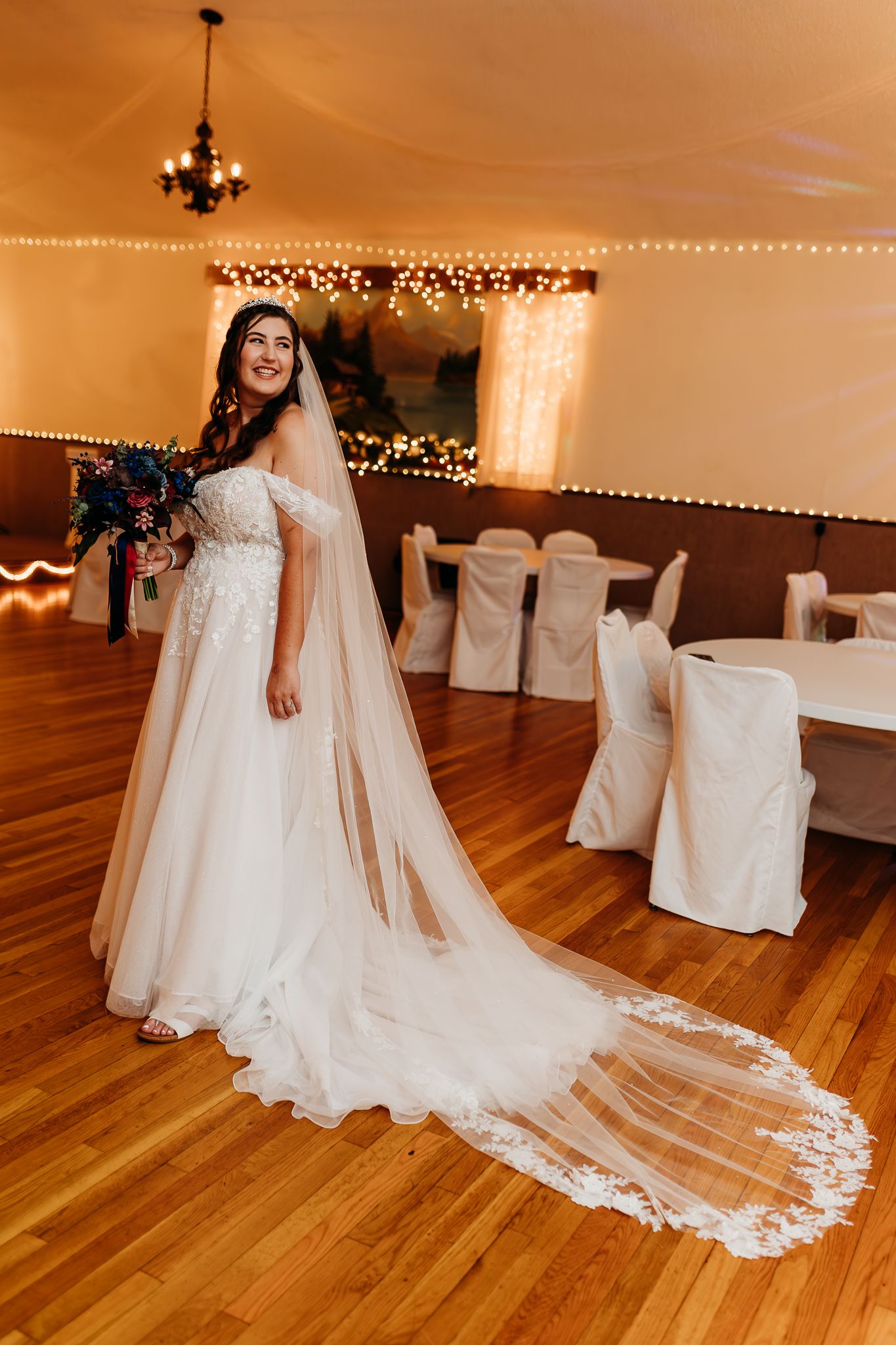 Smiling bride in off-shoulder lace gown with cathedral veil holds bouquet in warmly lit reception hall.
