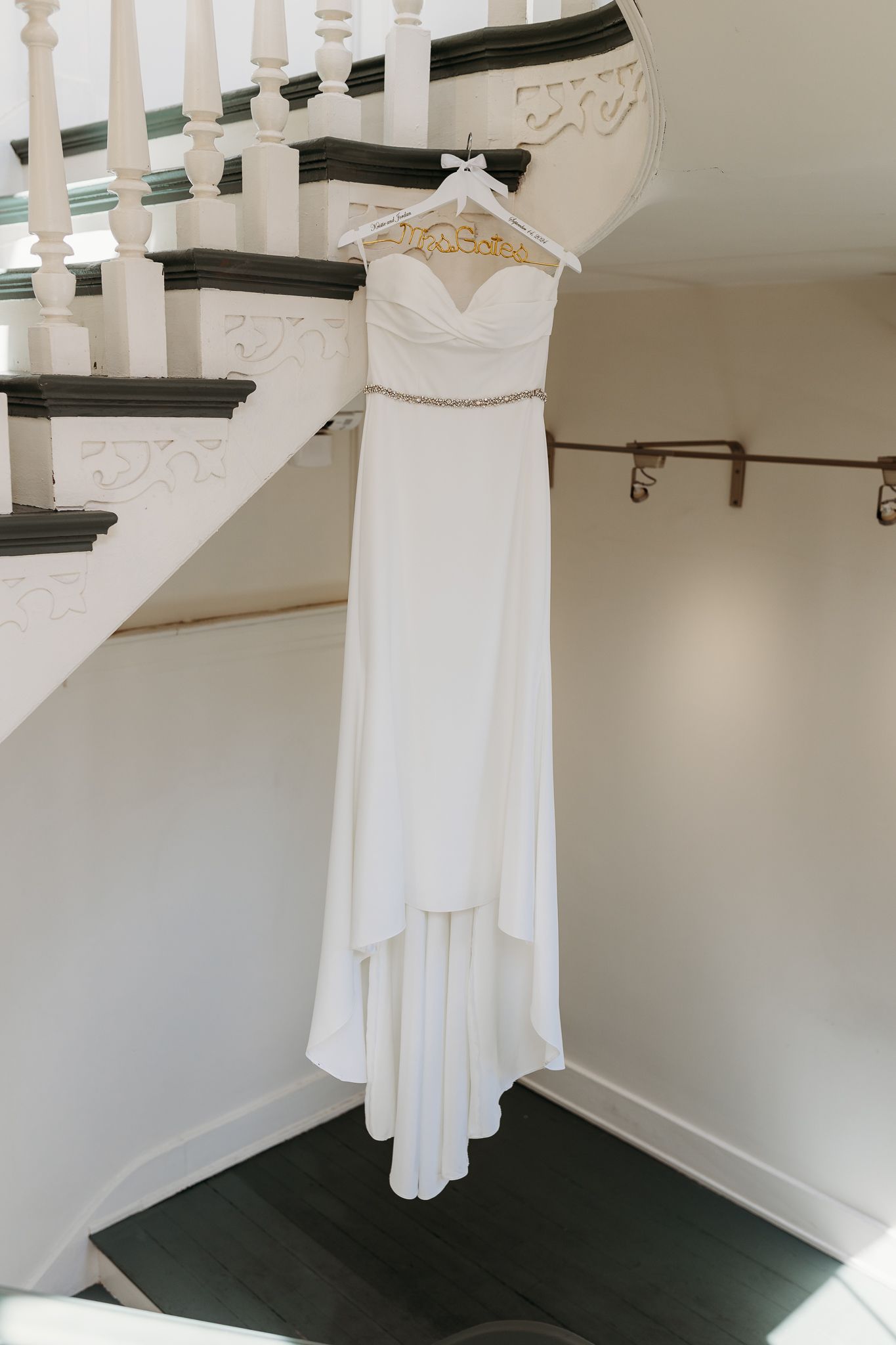 Simple strapless white wedding gown with beaded belt hanging on staircase banister.