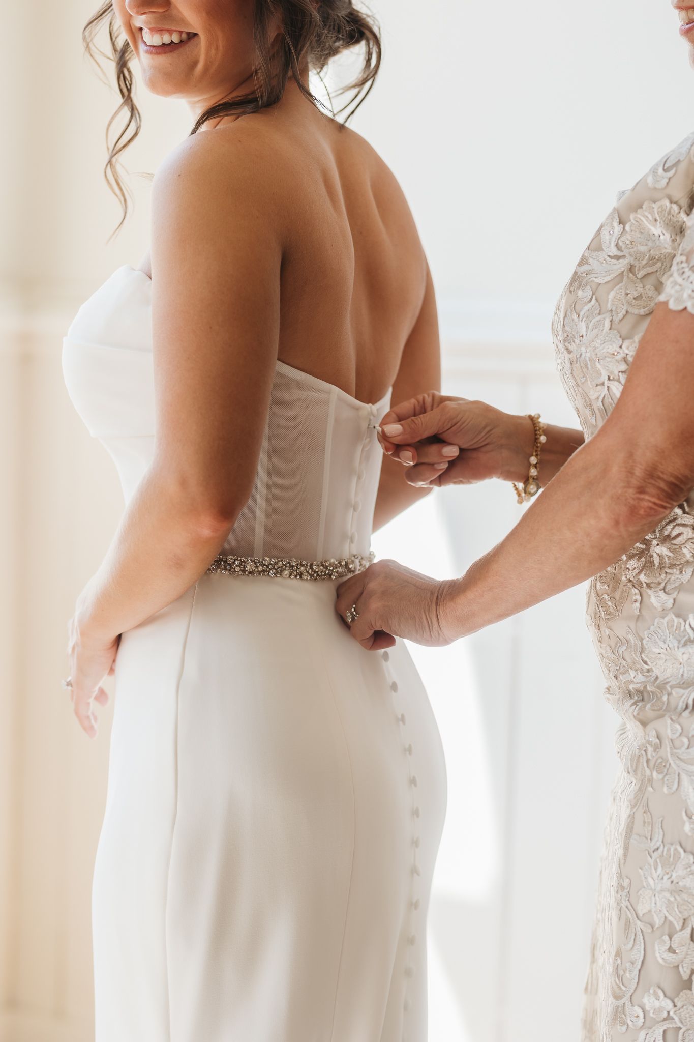 Mother fastening corset back of sleek strapless wedding dress with beaded belt on bride.