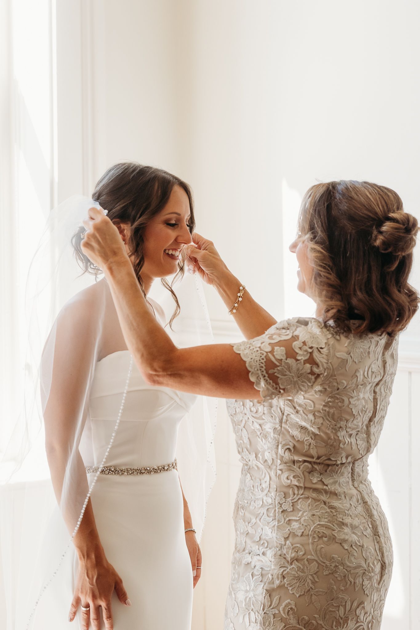 Mother adjusting veil on smiling bride in strapless gown with beaded sash by bright window.