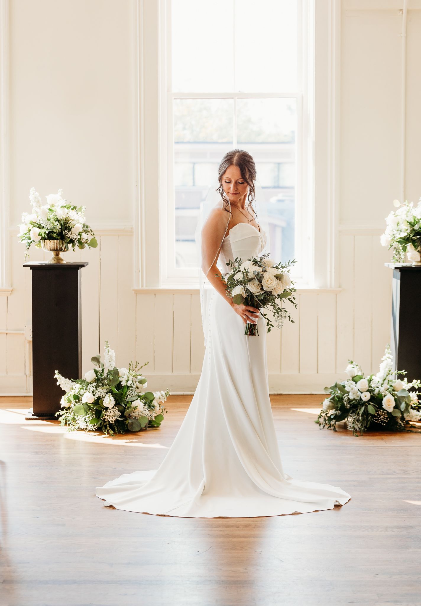 Bride in strapless satin gown holding bouquet surrounded by lush white floral arrangements indoors.