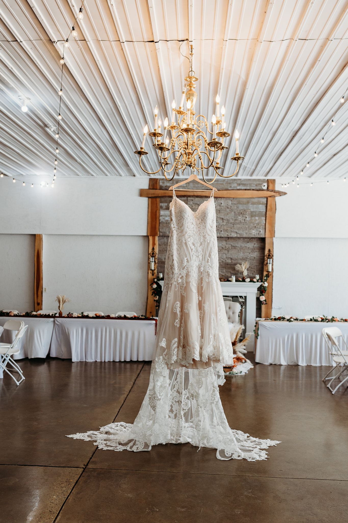 Lace wedding dress hanging from a chandelier in a rustic reception venue with string lights.