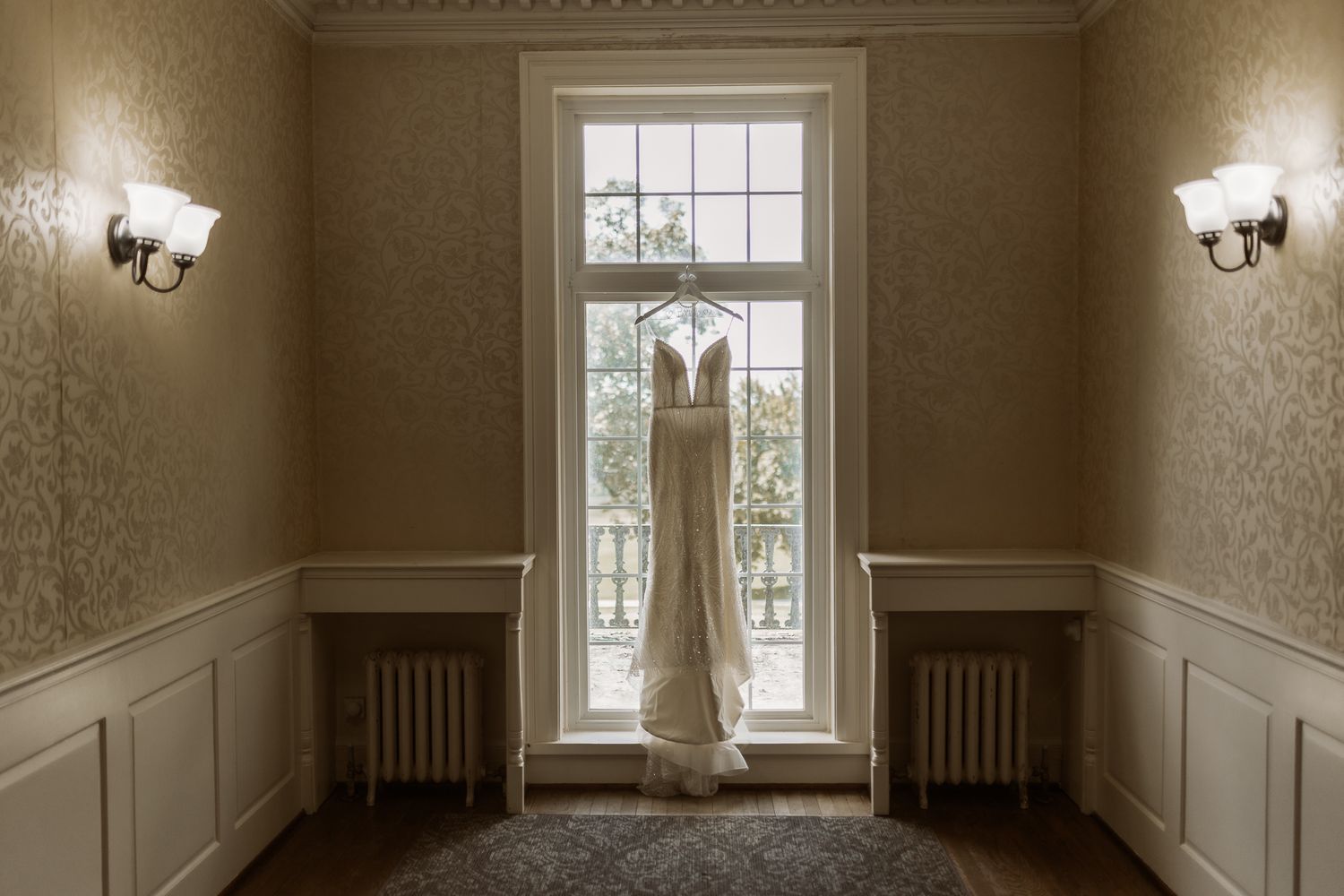 Elegant lace wedding dress hanging in a window of a classic room with ornate wallpaper.