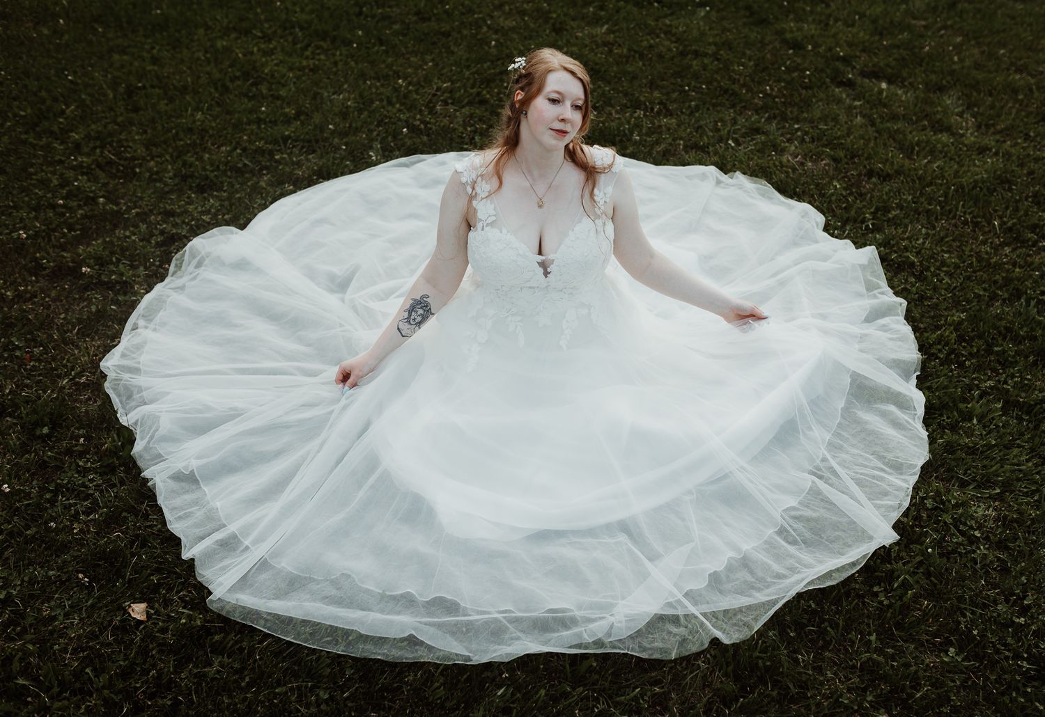 Bride in flowing white ballgown with lace bodice spreads skirt while seated on green grass.