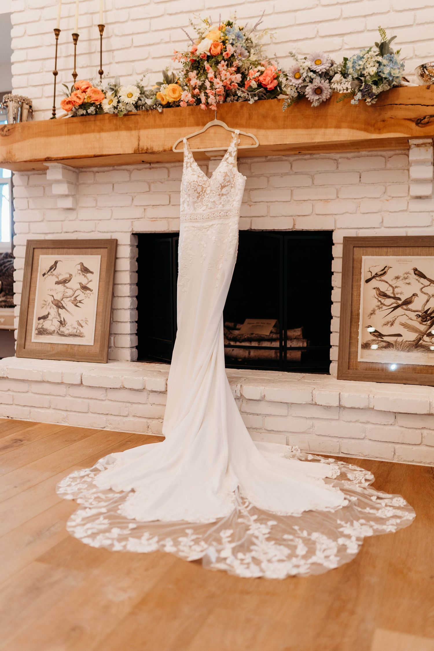 Fitted lace wedding dress with floral train displayed on hanger over white brick fireplace with florals.