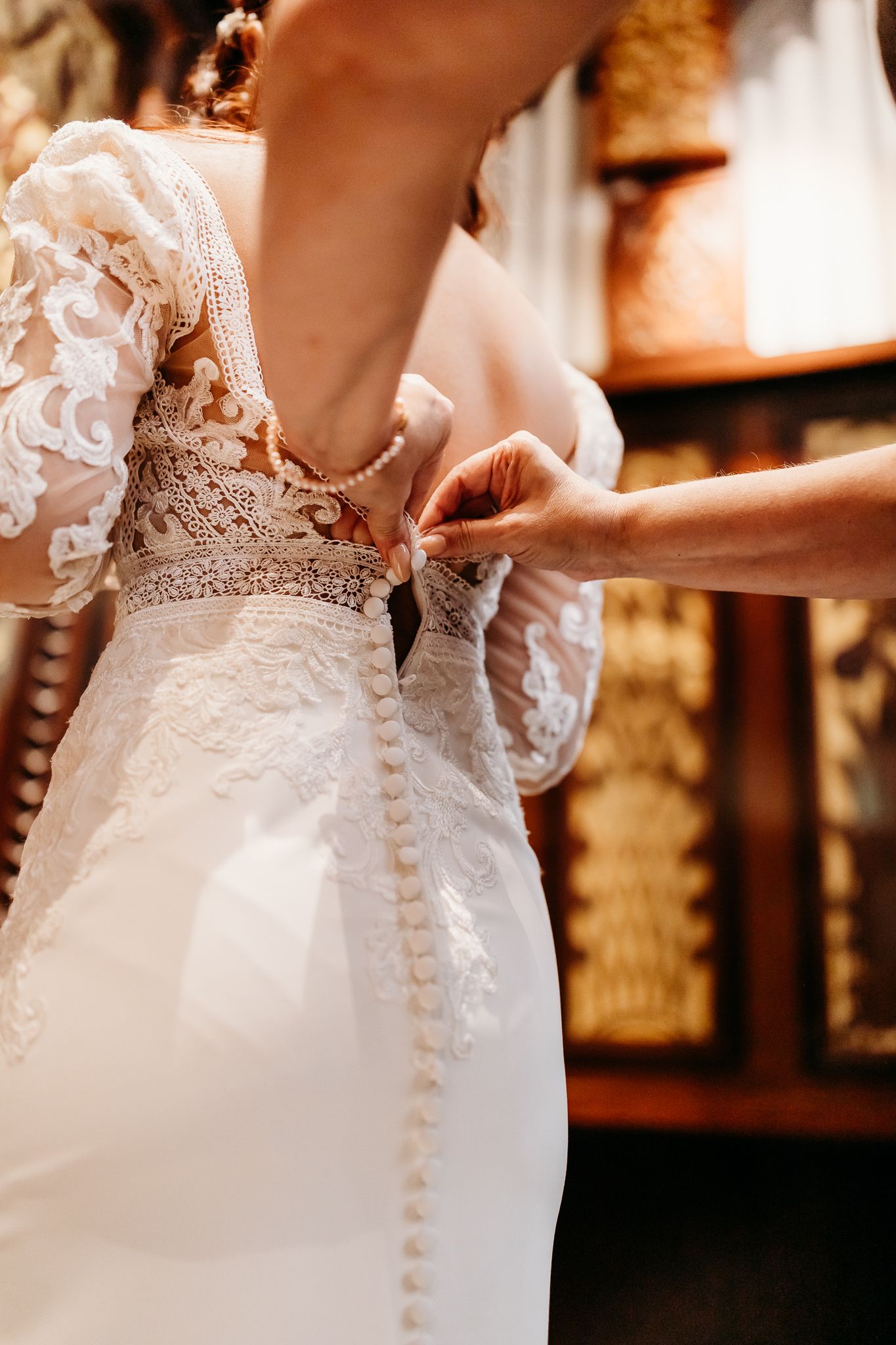 Bridesmaid buttoning the pearl back of a lace wedding gown on the bride.
