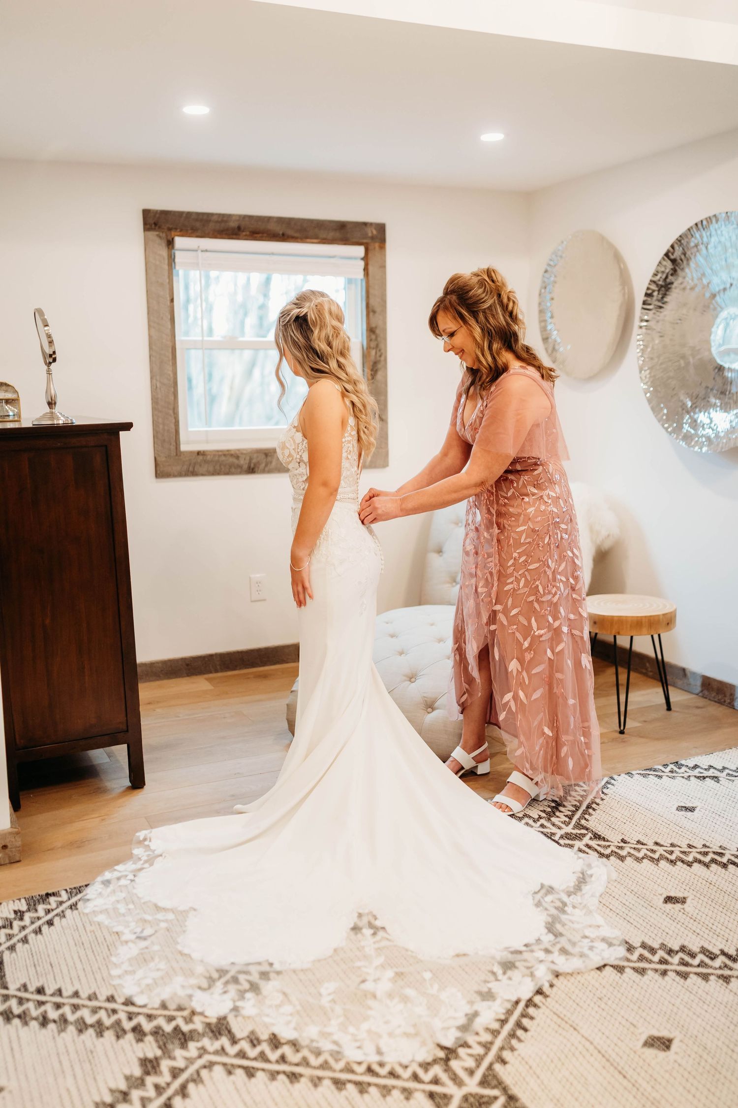 A bride in a white lace gown gets help fastening her dress from a woman in a pink beaded gown.