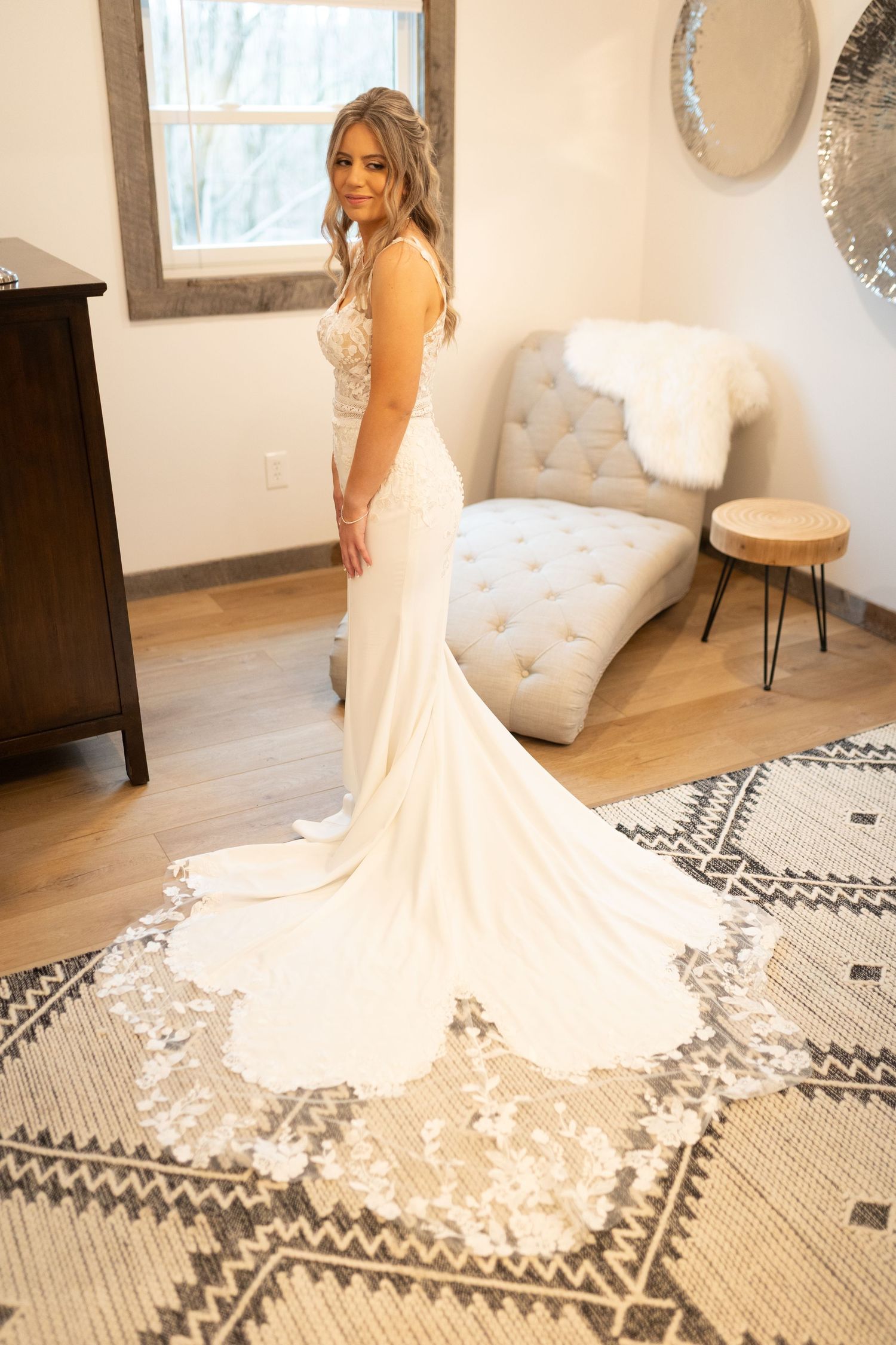 Elegant bride in a fitted white lace wedding gown with a floral train poses in a rustic bridal suite.