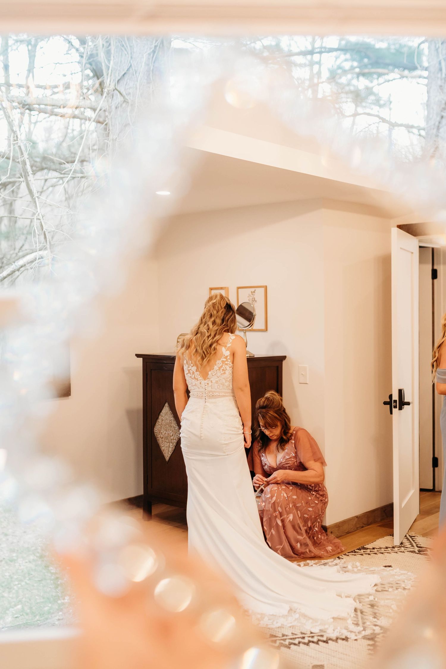 A helper kneels to adjust the train of a brides white lace gown in a warmly lit bridal suite.