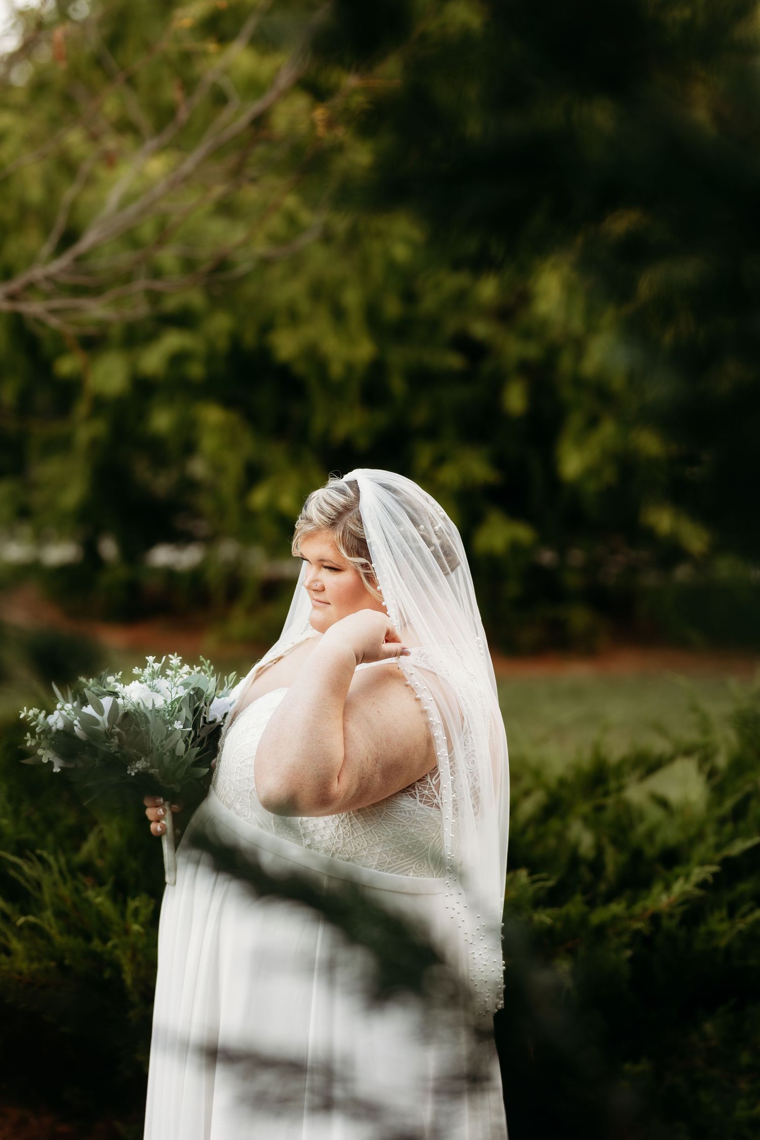 Plus-size bride in white lace gown holding bouquet while posing outdoors in green garden.