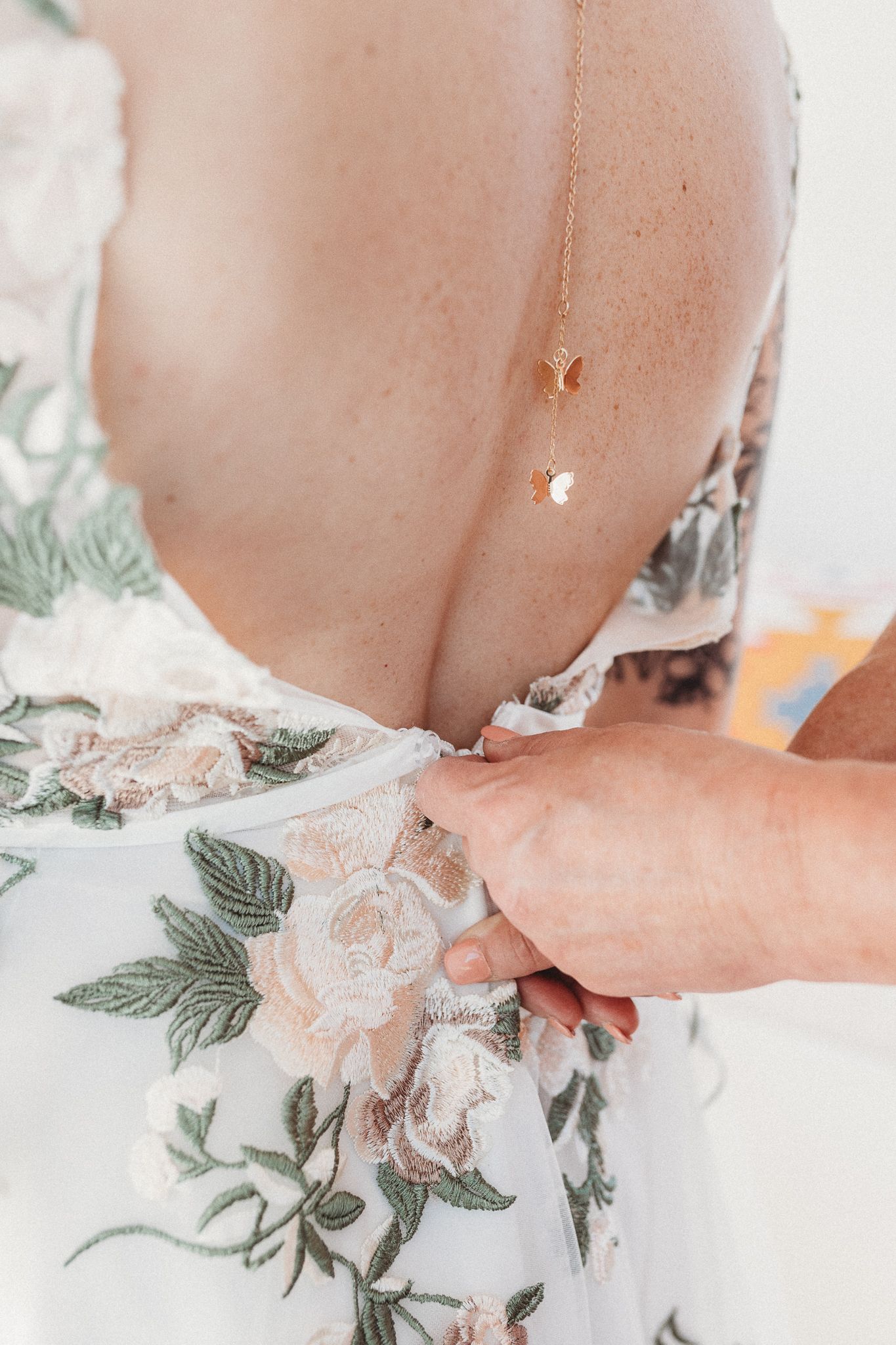 Close-up of floral embroidered wedding dress being fastened with delicate gold back necklace detail.