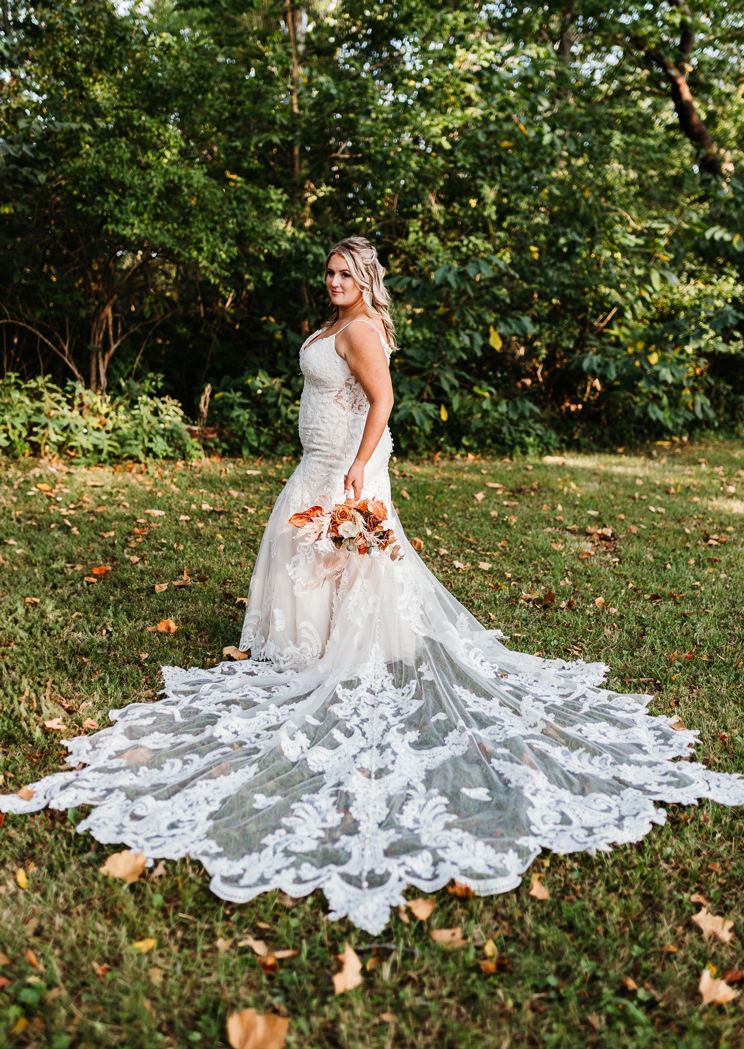Bride in a mermaid lace gown with cathedral train holding fall bouquet in a garden.