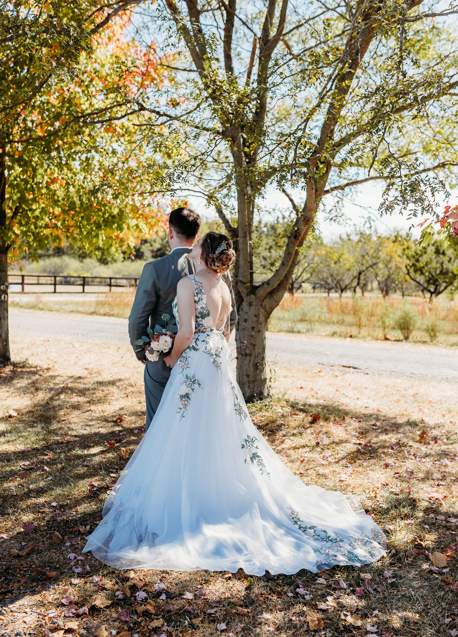 Bride and groom stand under autumn tree, showcasing backless floral wedding gown with long train.