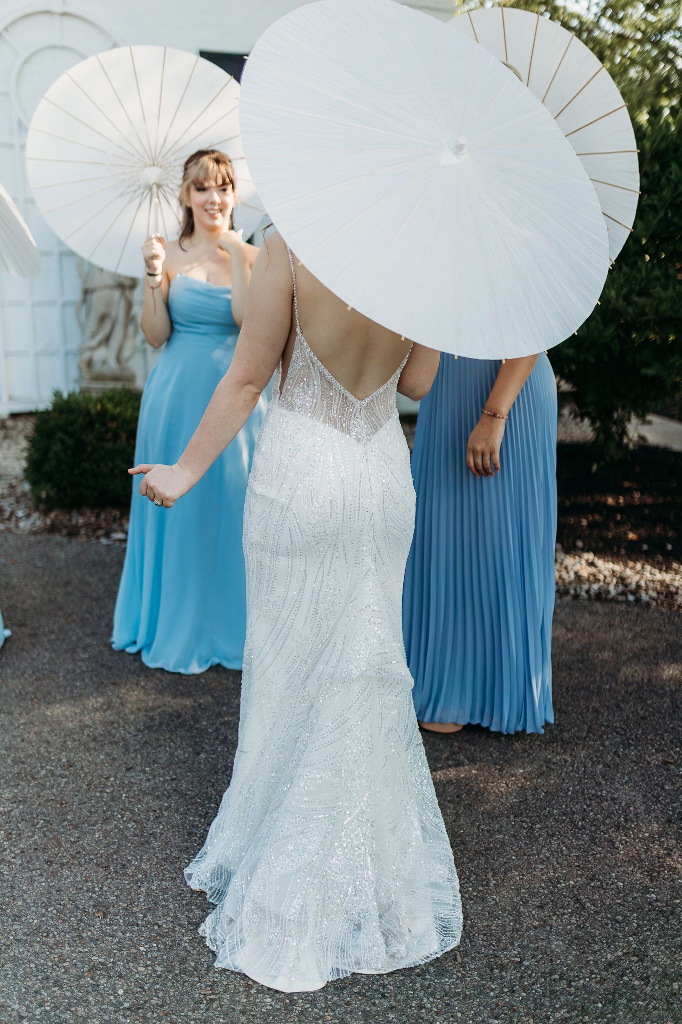 Bride in fitted lace gown with bridesmaids in blue dresses holding white parasols outdoors.