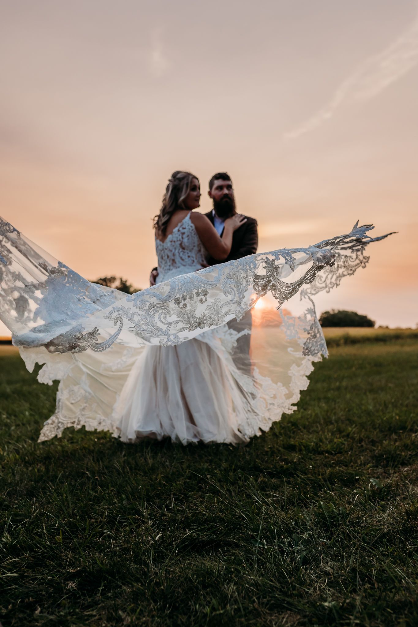 Bride and groom embrace at sunset as her lace veil dramatically billows in the wind.