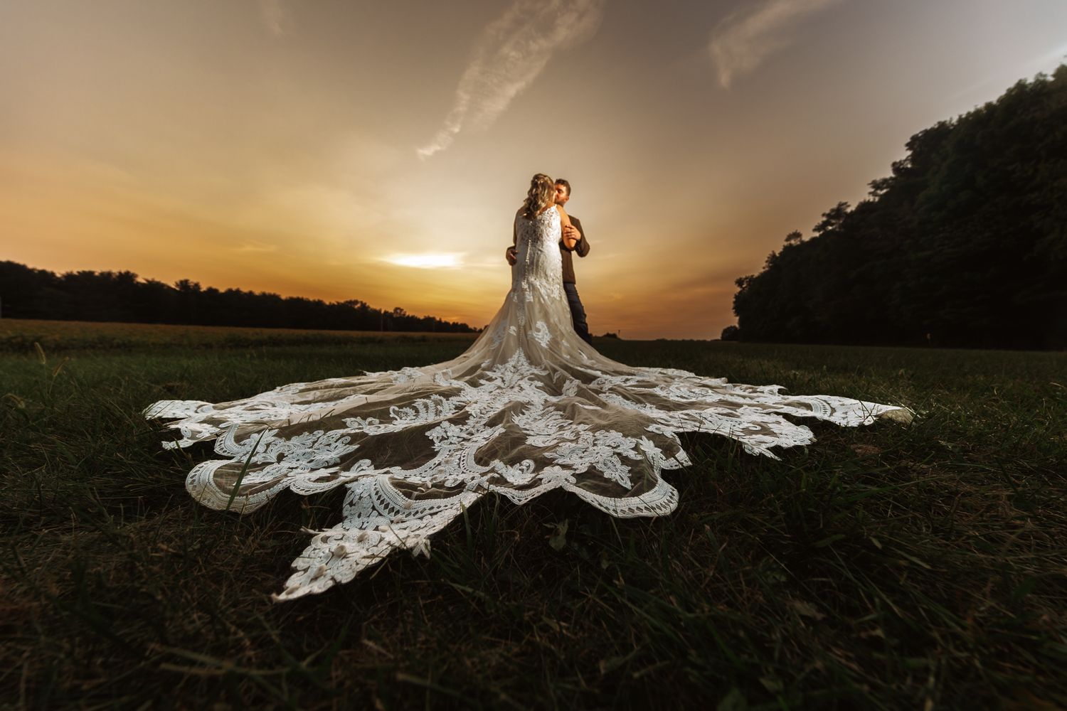 Bride and groom embrace at sunset with stunning cathedral lace train spread across green field.