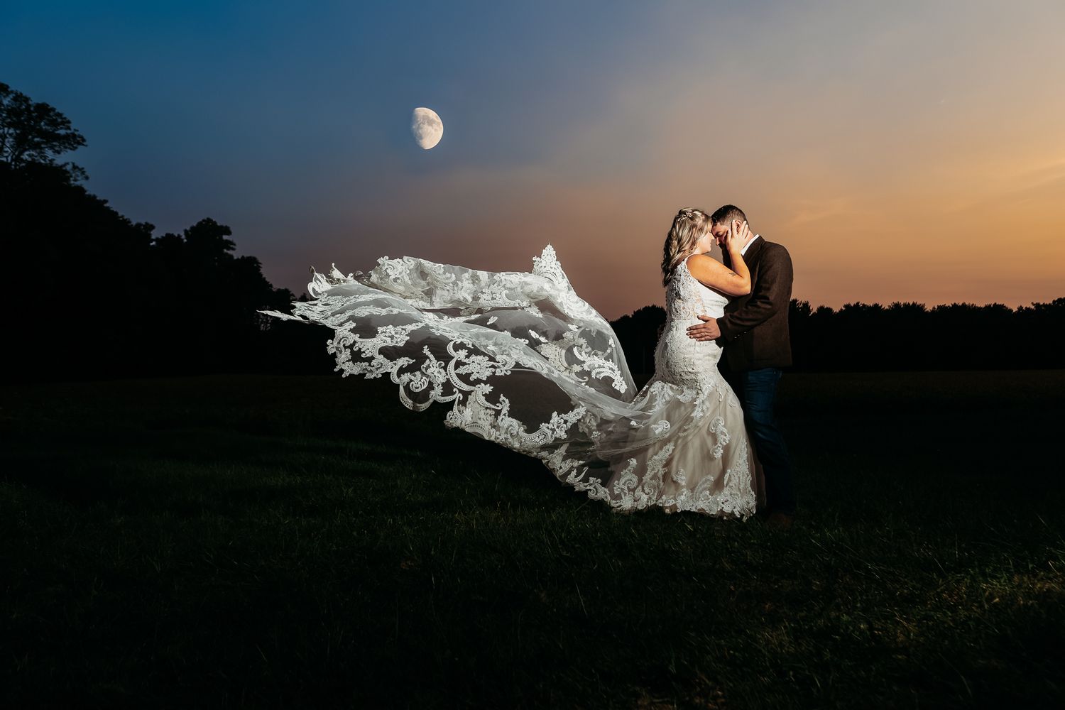 Bride and groom share a kiss under a crescent moon with a flowing lace veil at dusk.