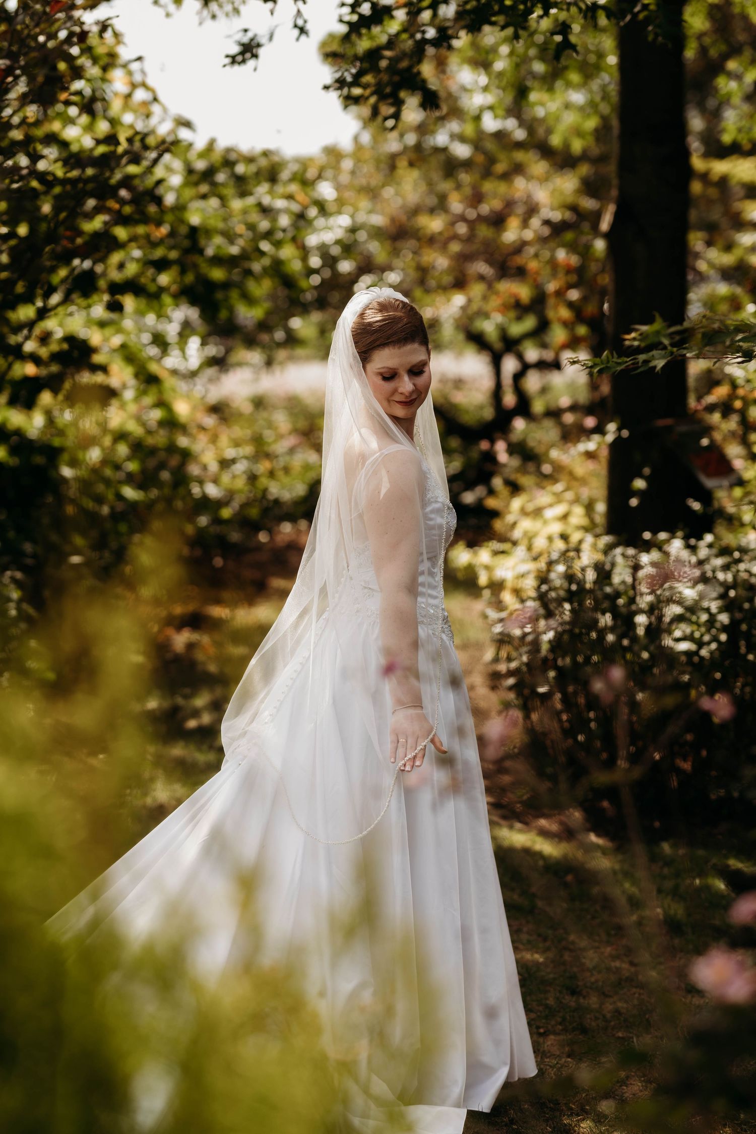 Bride in white gown with sheer veil poses gracefully in sunlit garden surrounded by lush greenery.