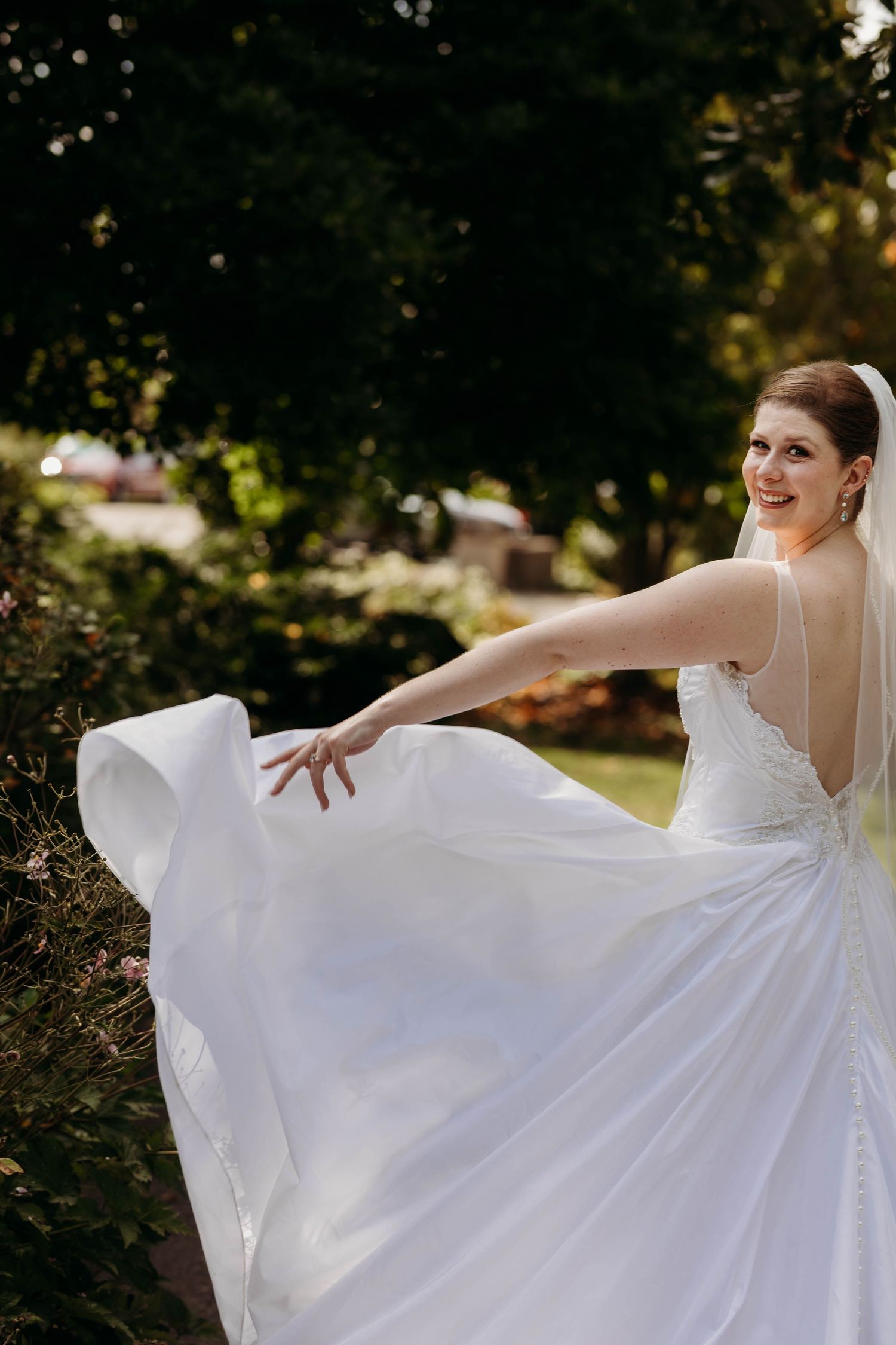 Smiling bride twirls white wedding gown with long flowing train in sunlit outdoor garden setting.