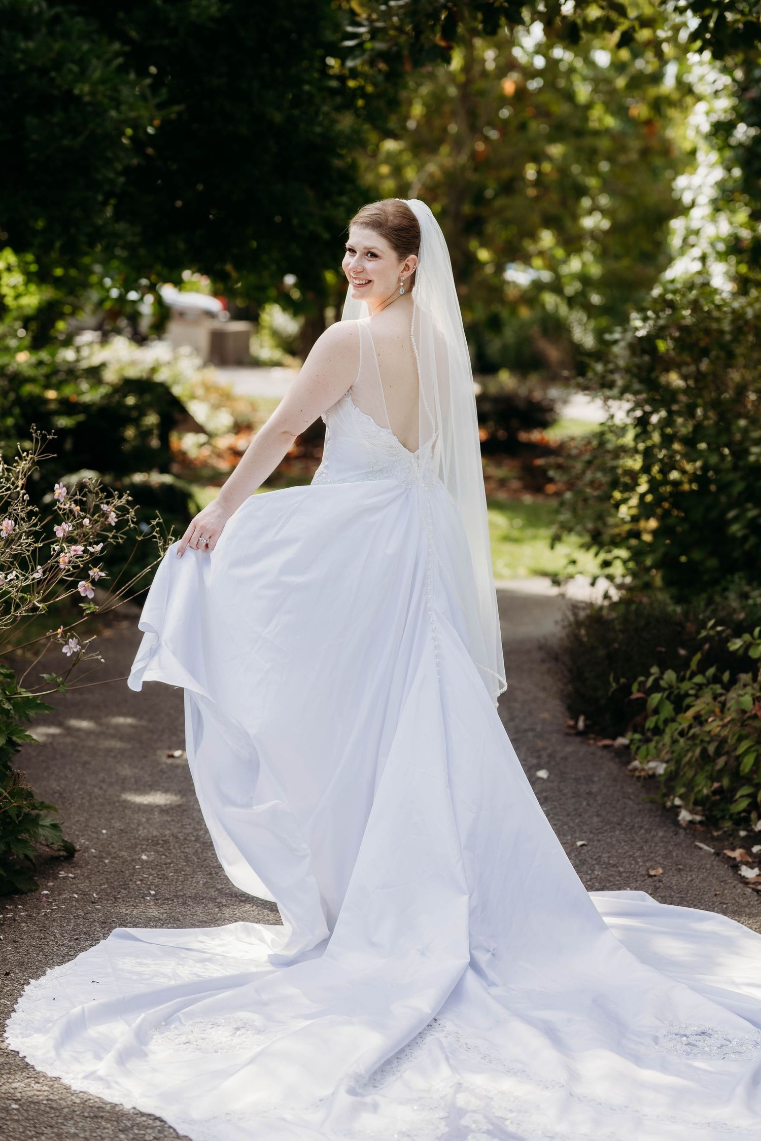 Bride in sleeveless backless gown with cathedral train and veil poses on garden path in sunlight.