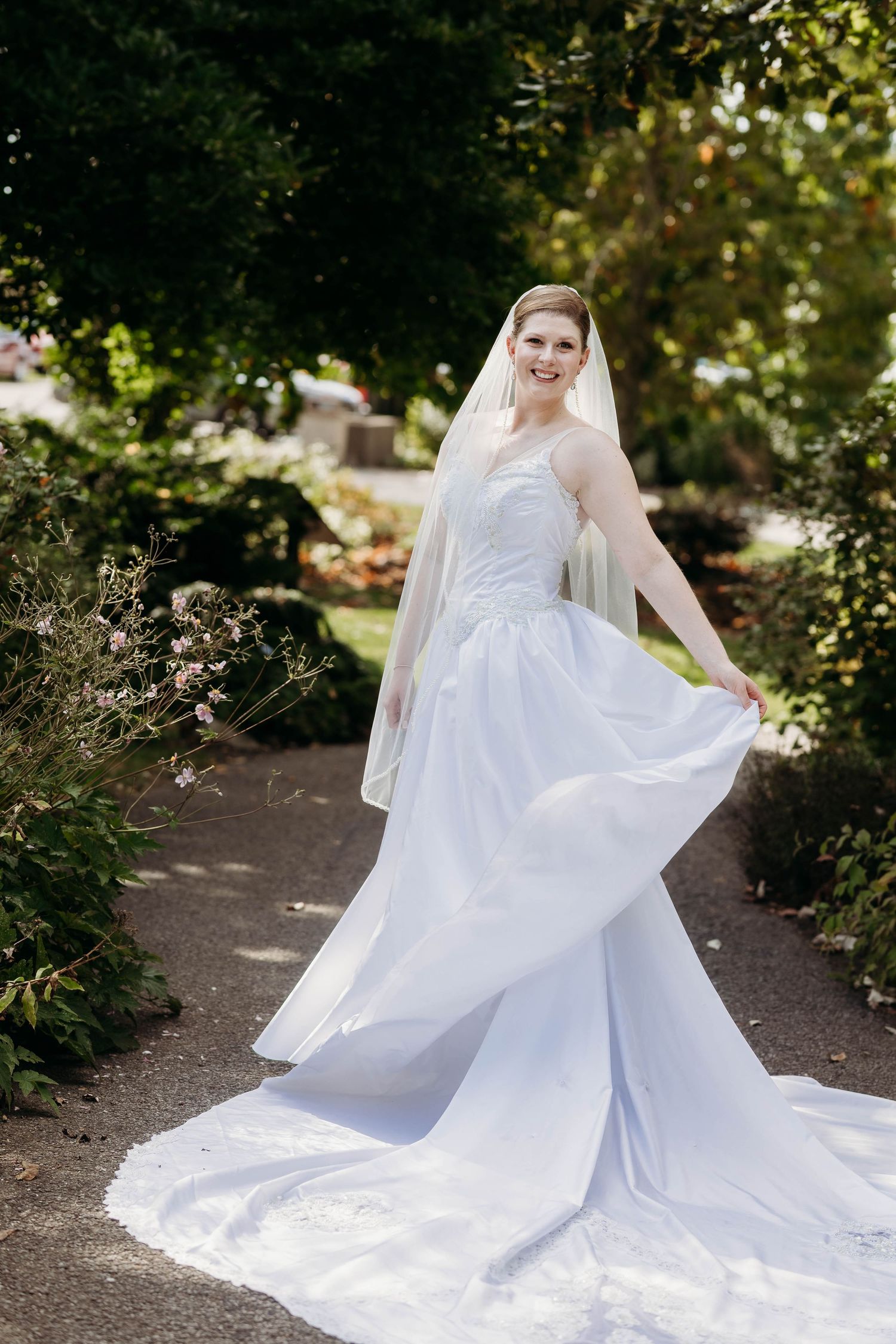 Bride in white ball gown and veil twirling on garden path surrounded by lush greenery.