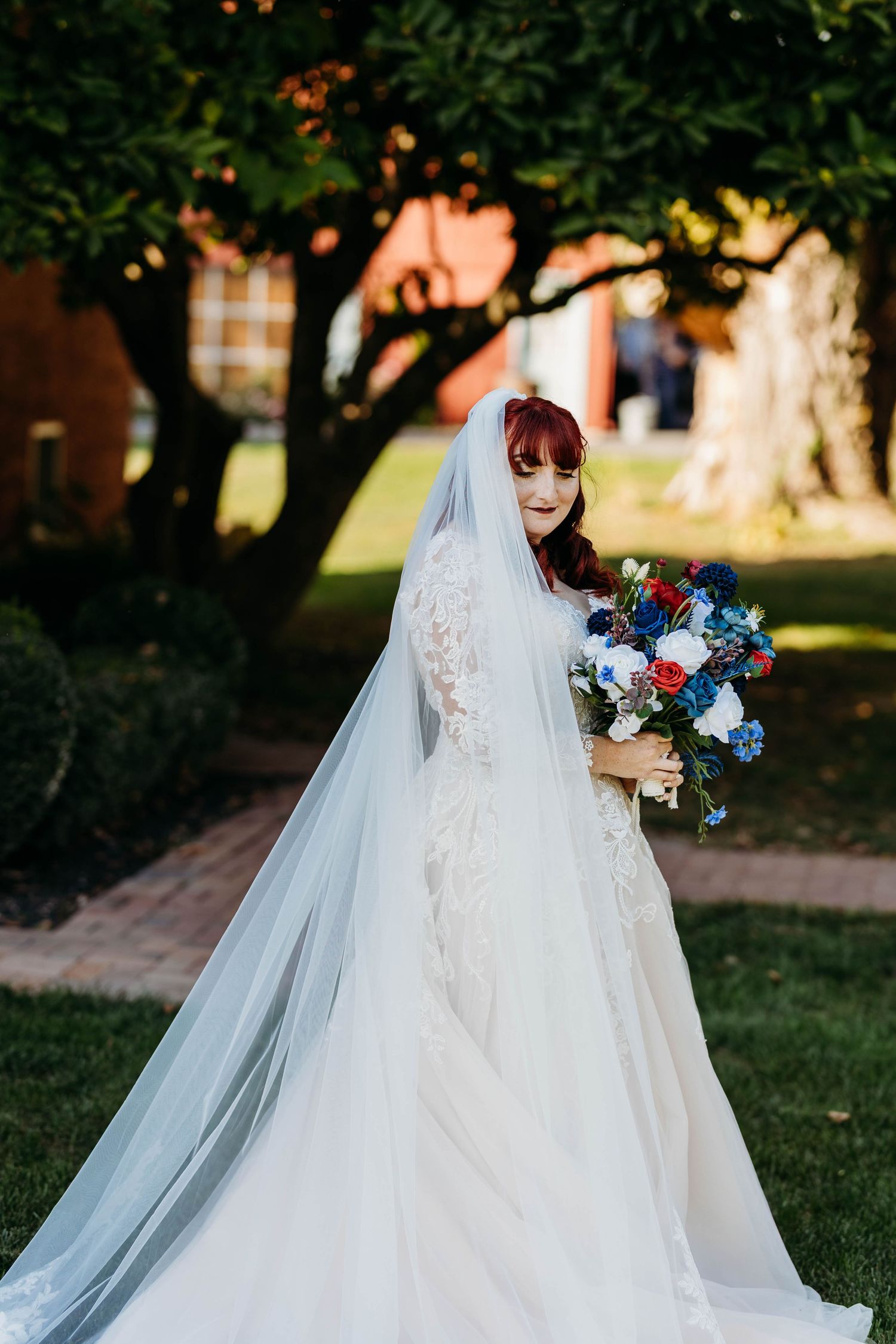 Red-haired bride in lace wedding gown with veil holding blue and red bouquet outdoors.