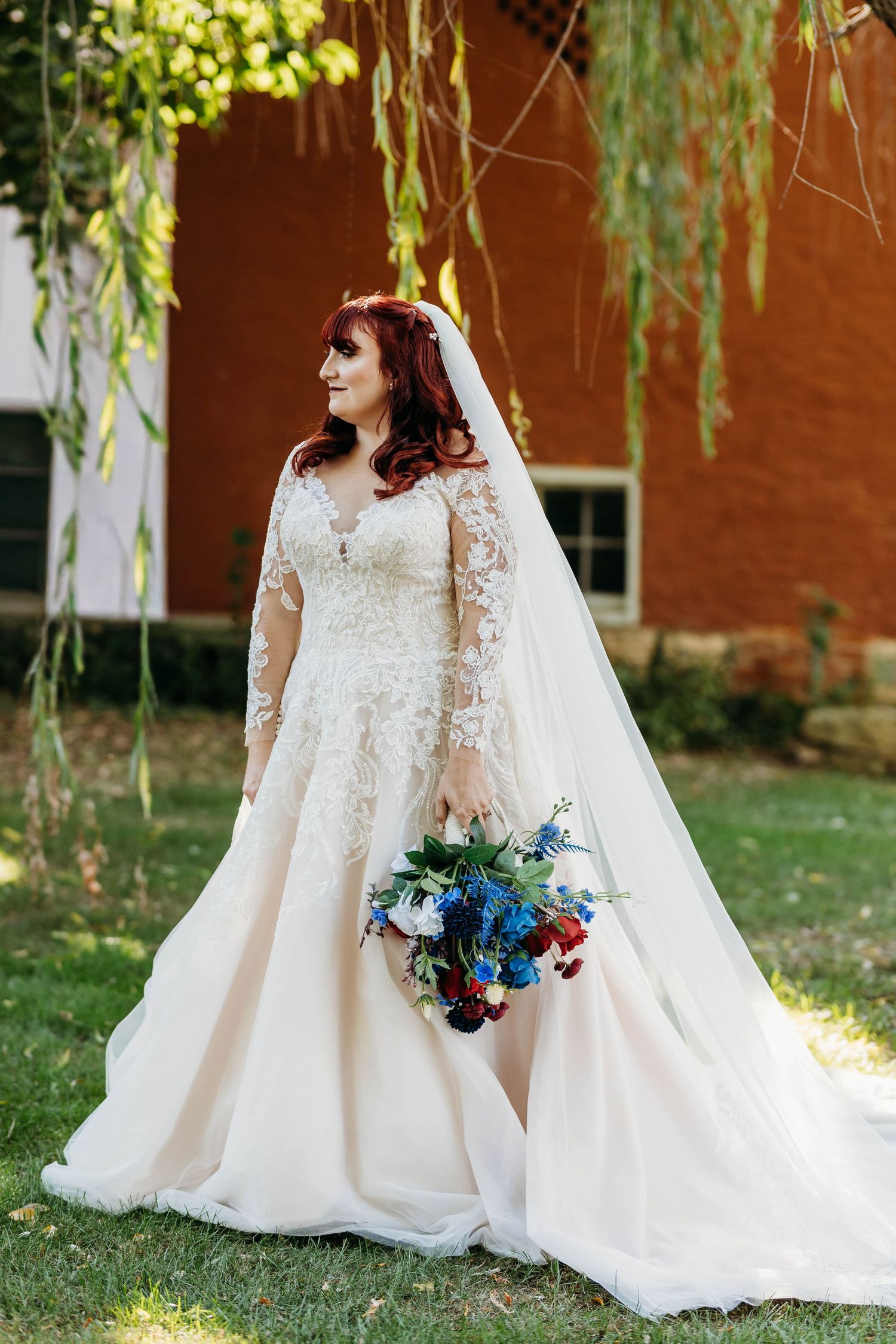 Bride in long-sleeve lace wedding gown holding colorful bouquet beneath weeping willow tree.