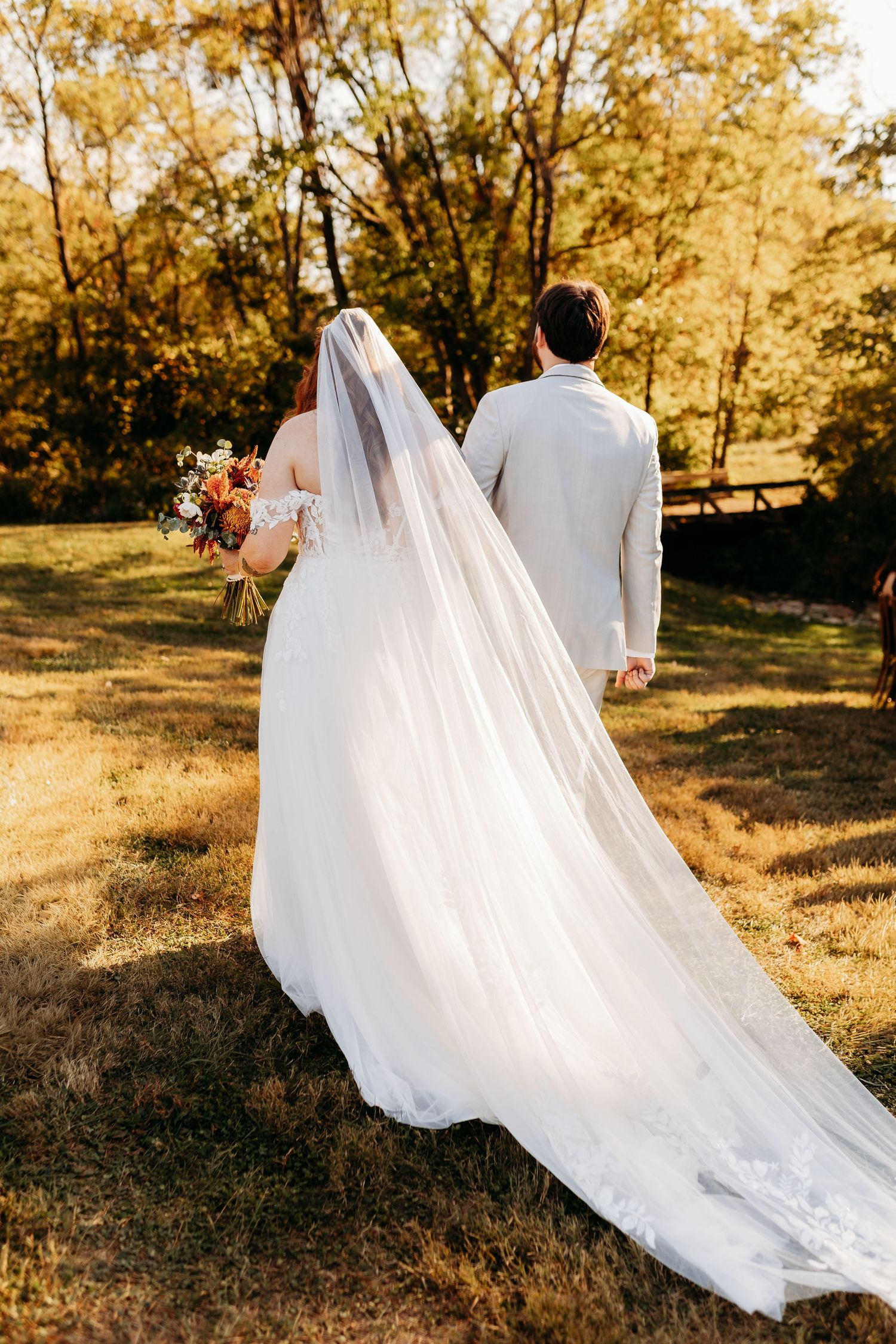Bride and groom walking hand-in-hand through golden autumn field, veil trailing behind.